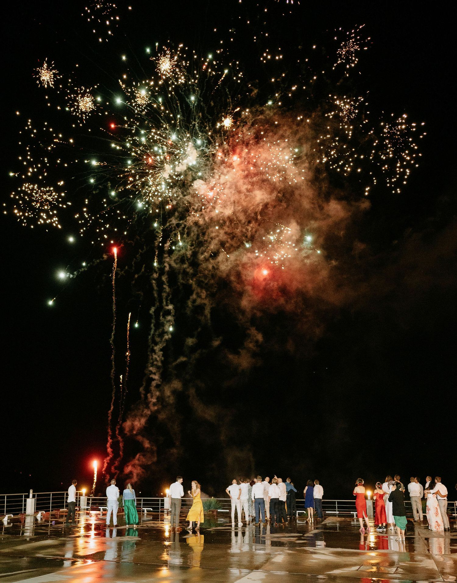 Nighttime wedding celebration with colorful fireworks display illuminating guests on an outdoor deck.