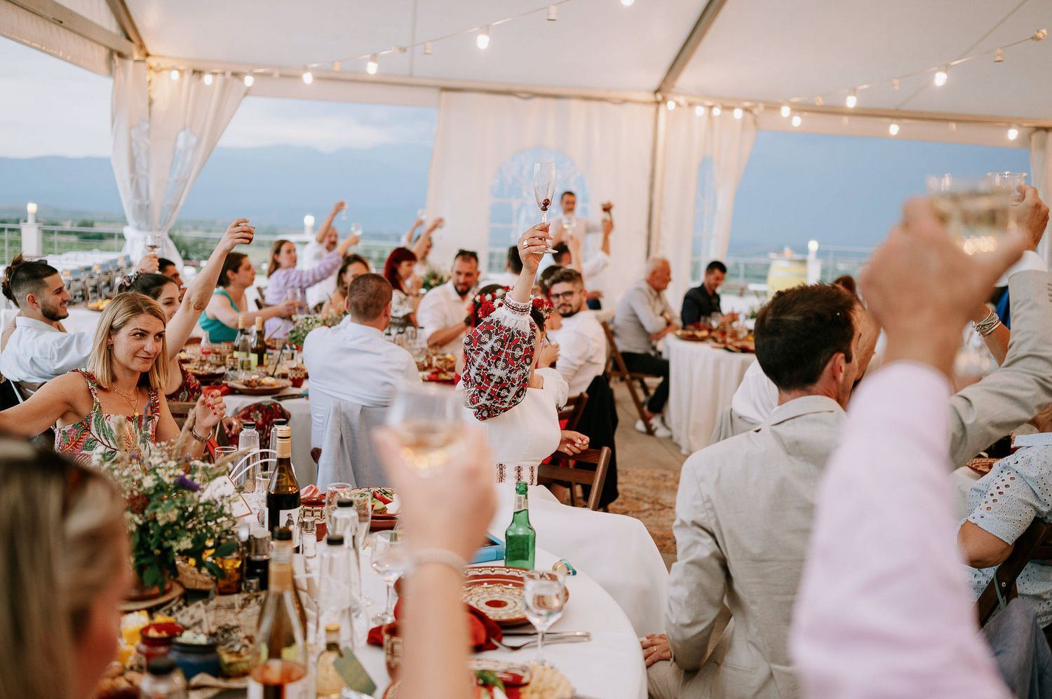 Wedding guests cheering with raised glasses during reception toasts under white tent canopy.