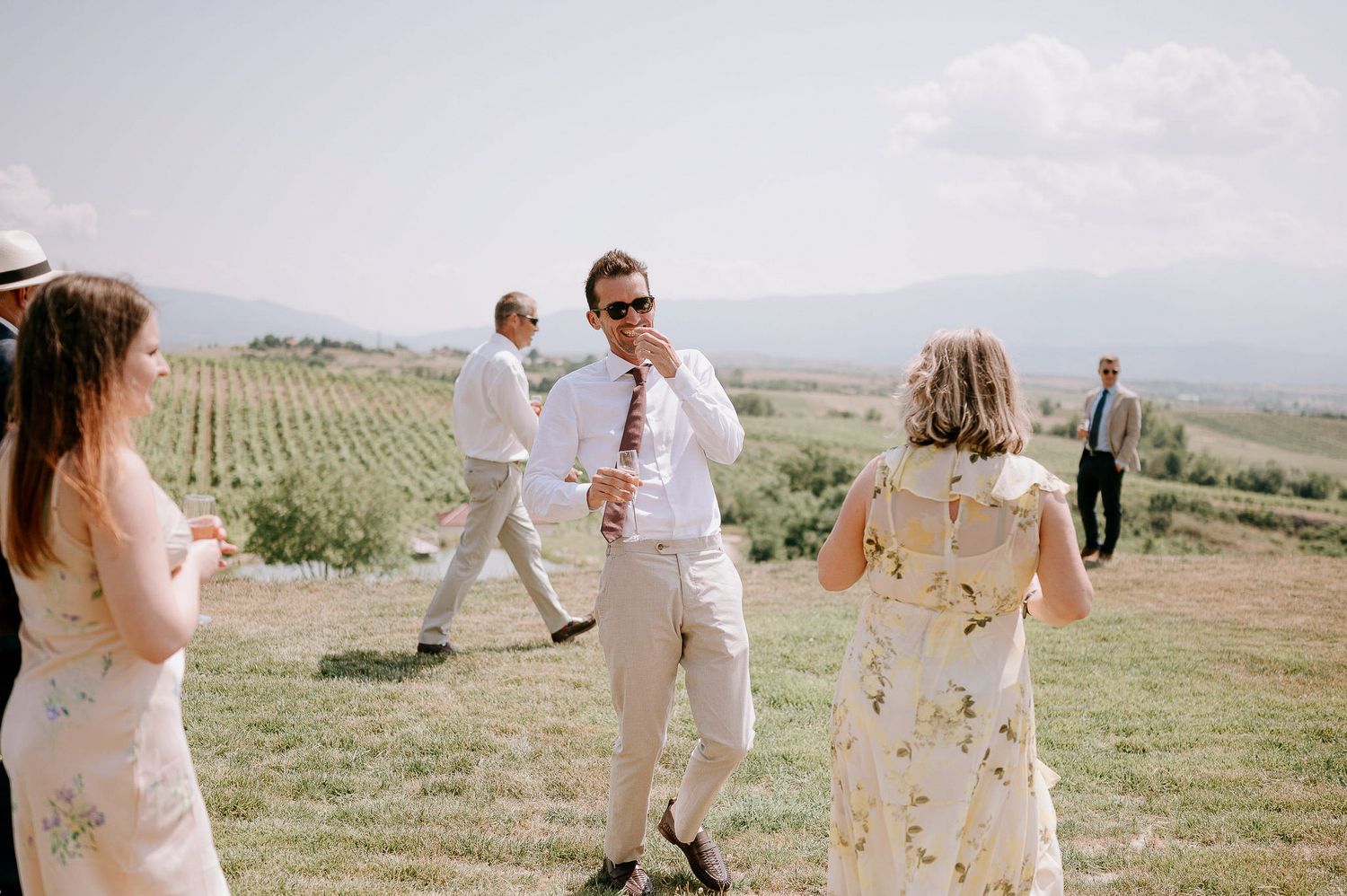 Guests in light summer attire gather on a hilltop overlooking vineyards on a sunny afternoon.