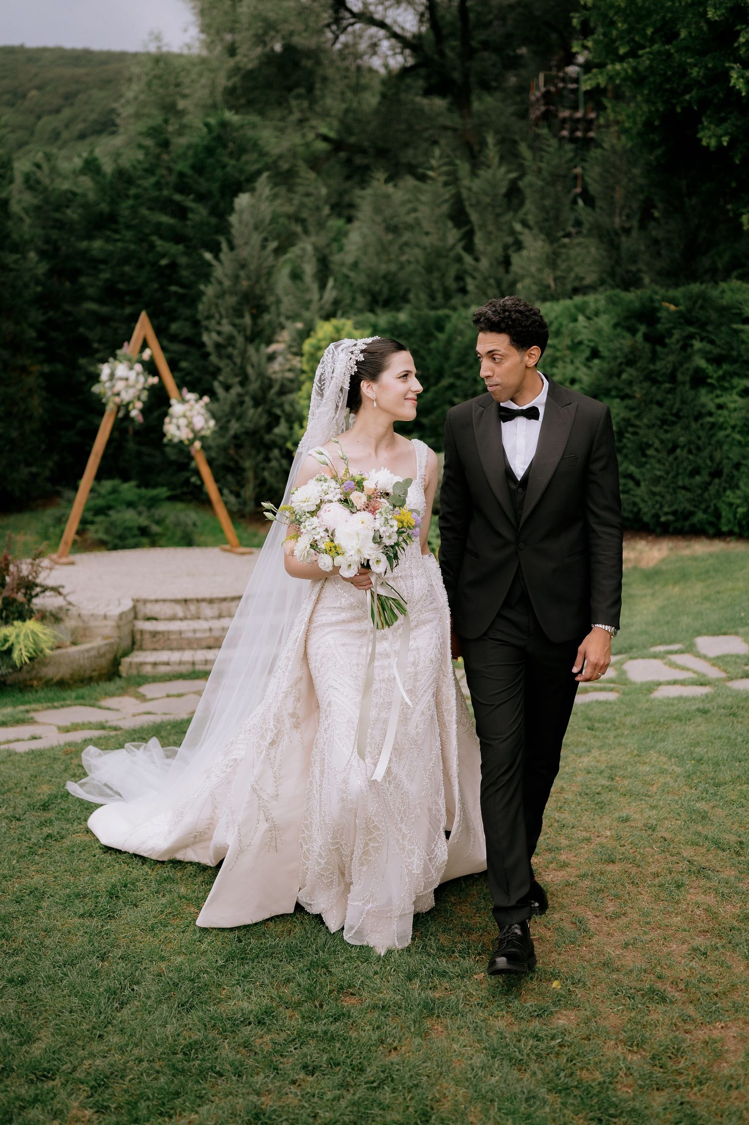 Bride in flowing gown and groom in tuxedo walking through garden setting.