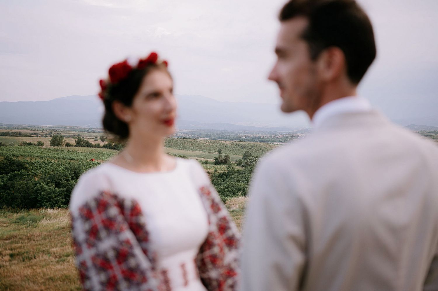 A wedding couple poses on a hillside with embroidered folk dress details and scenic mountain views.