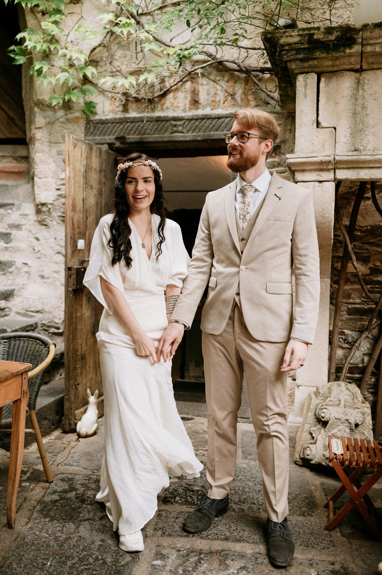 Couple poses casually against stone wall in coordinated beige and white outfits.