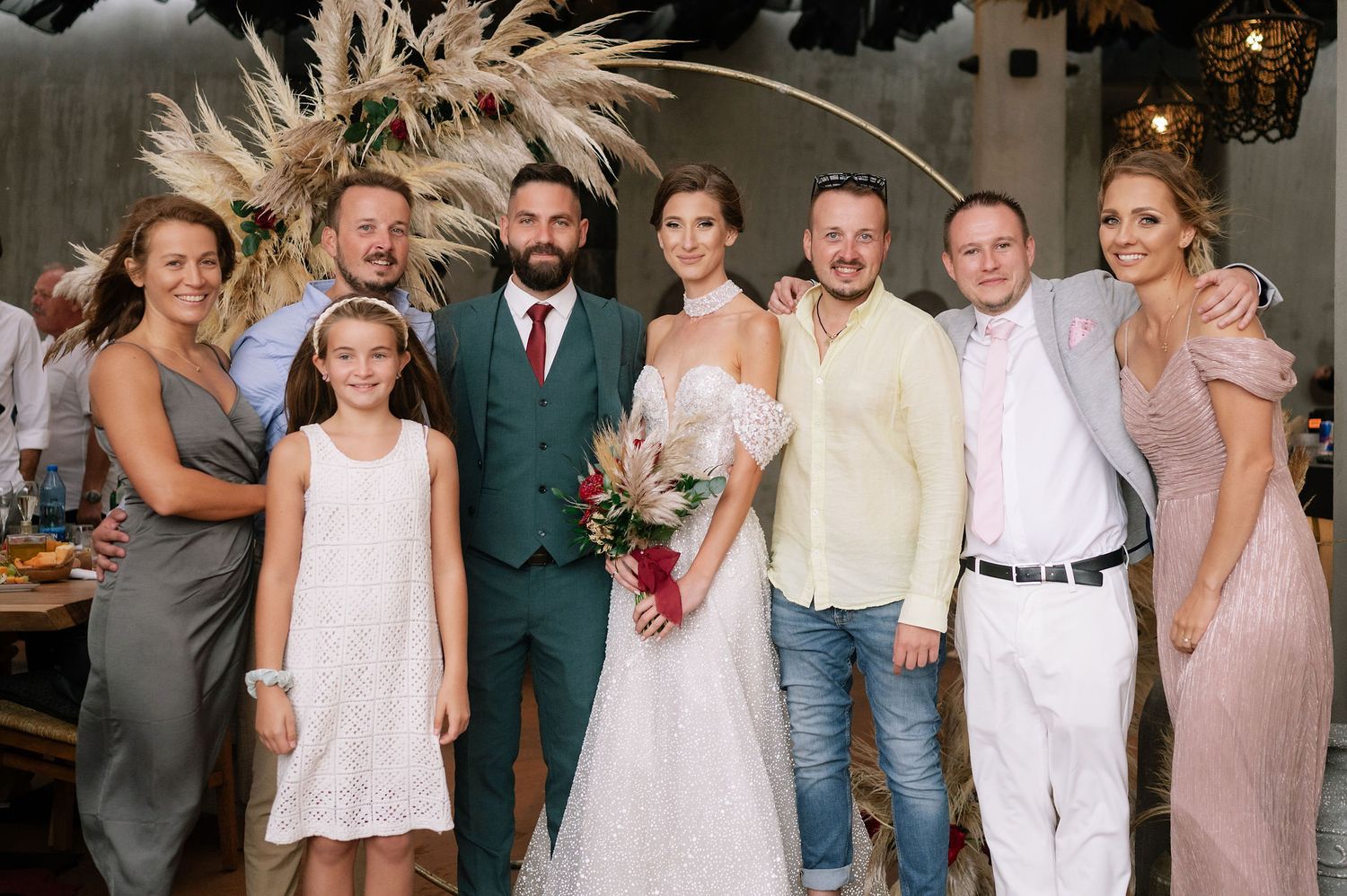 Wedding party group photo featuring a mix of formal and casual attire with pampas grass decor in the background.