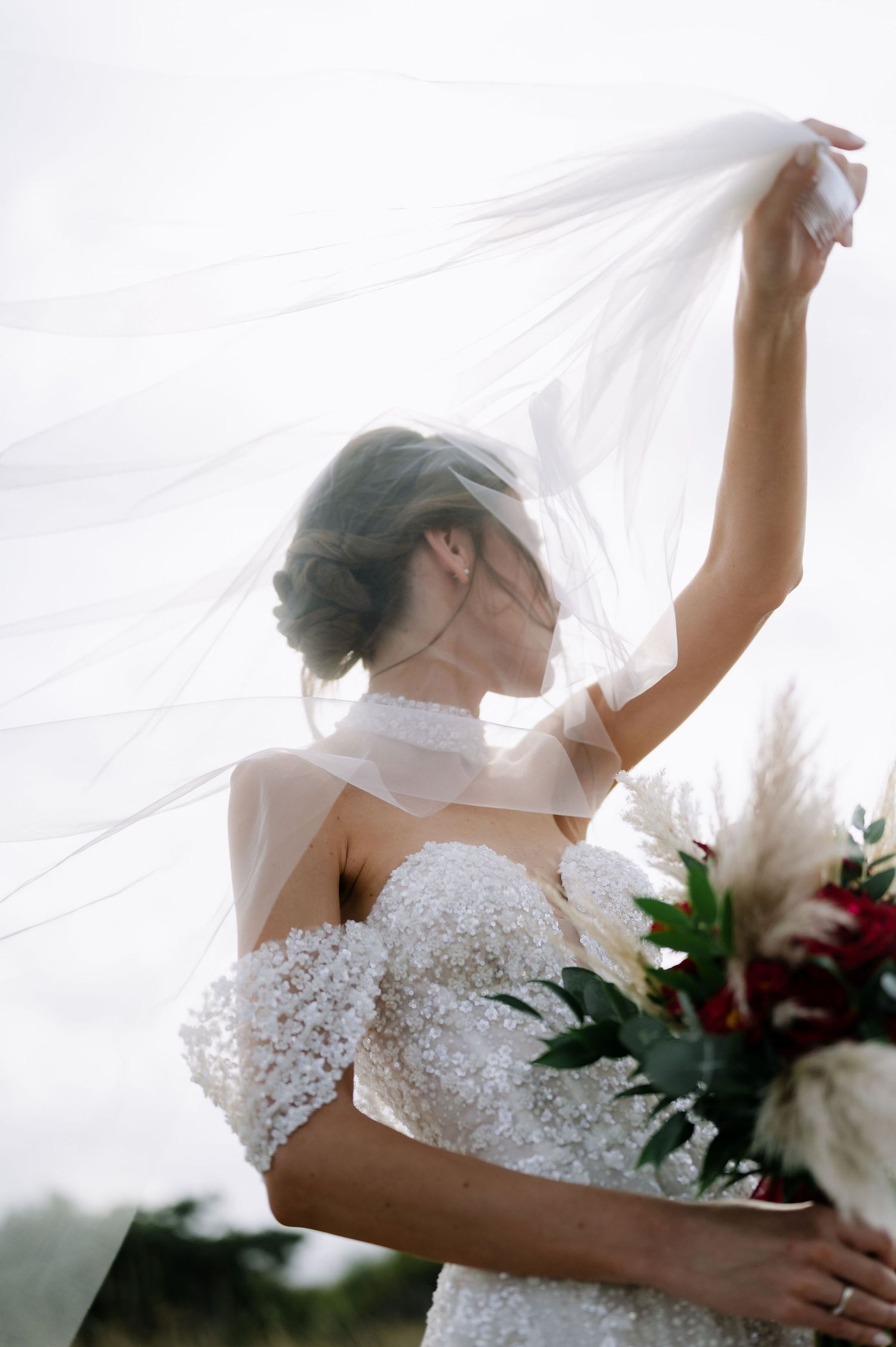 Artistic backlit portrait of a bride with flowing veil holding a dramatic red and white bouquet.