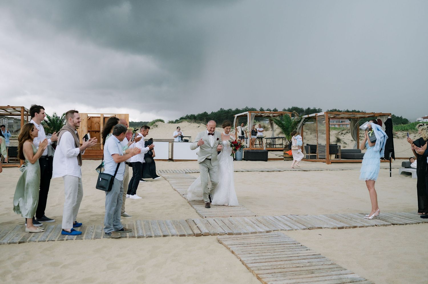 Panoramic view of beach wedding setup with wooden walkway and guests in formal wear.