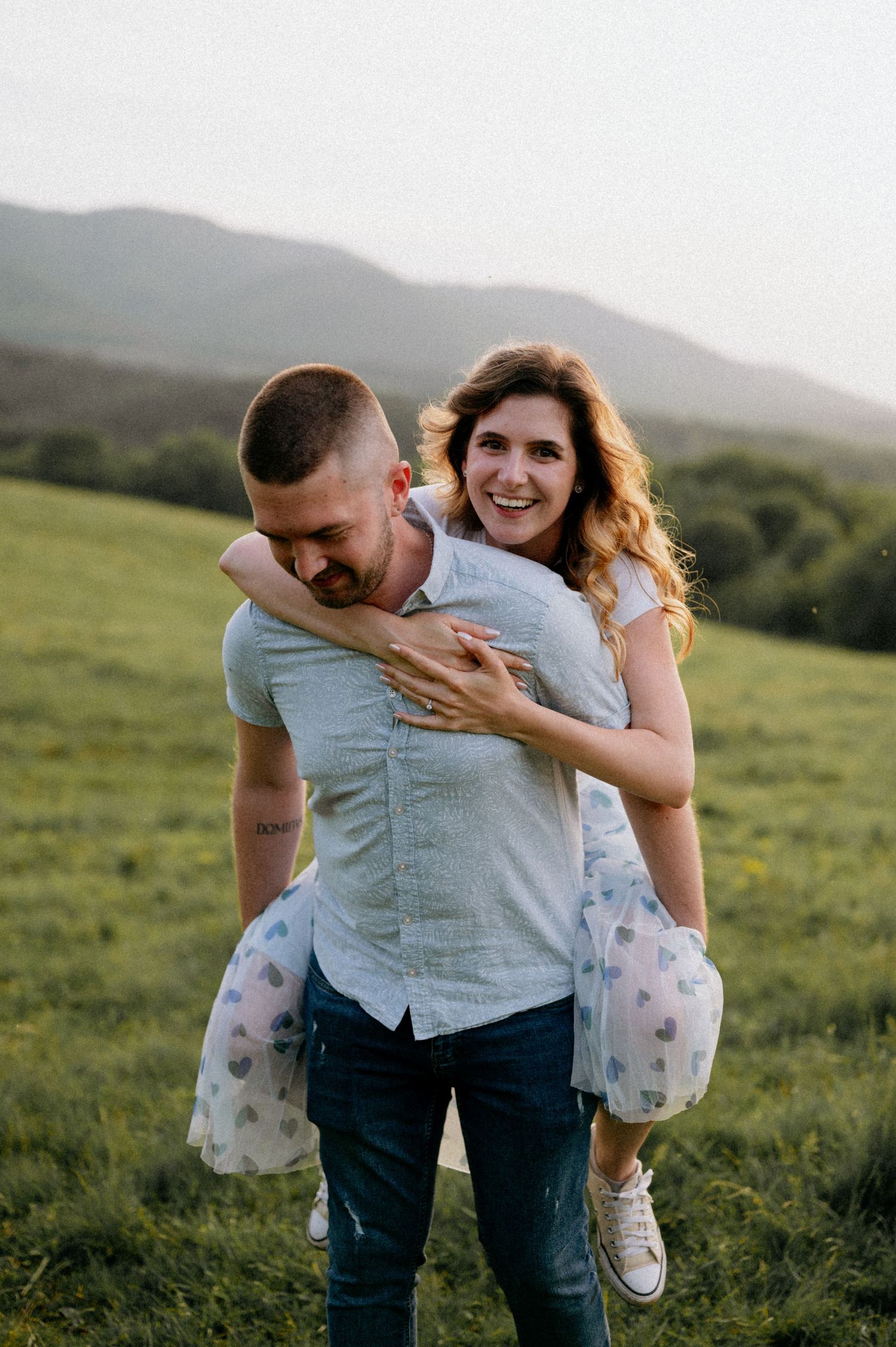 A playful couple shares a joyful moment together on a grassy hillside at sunset.