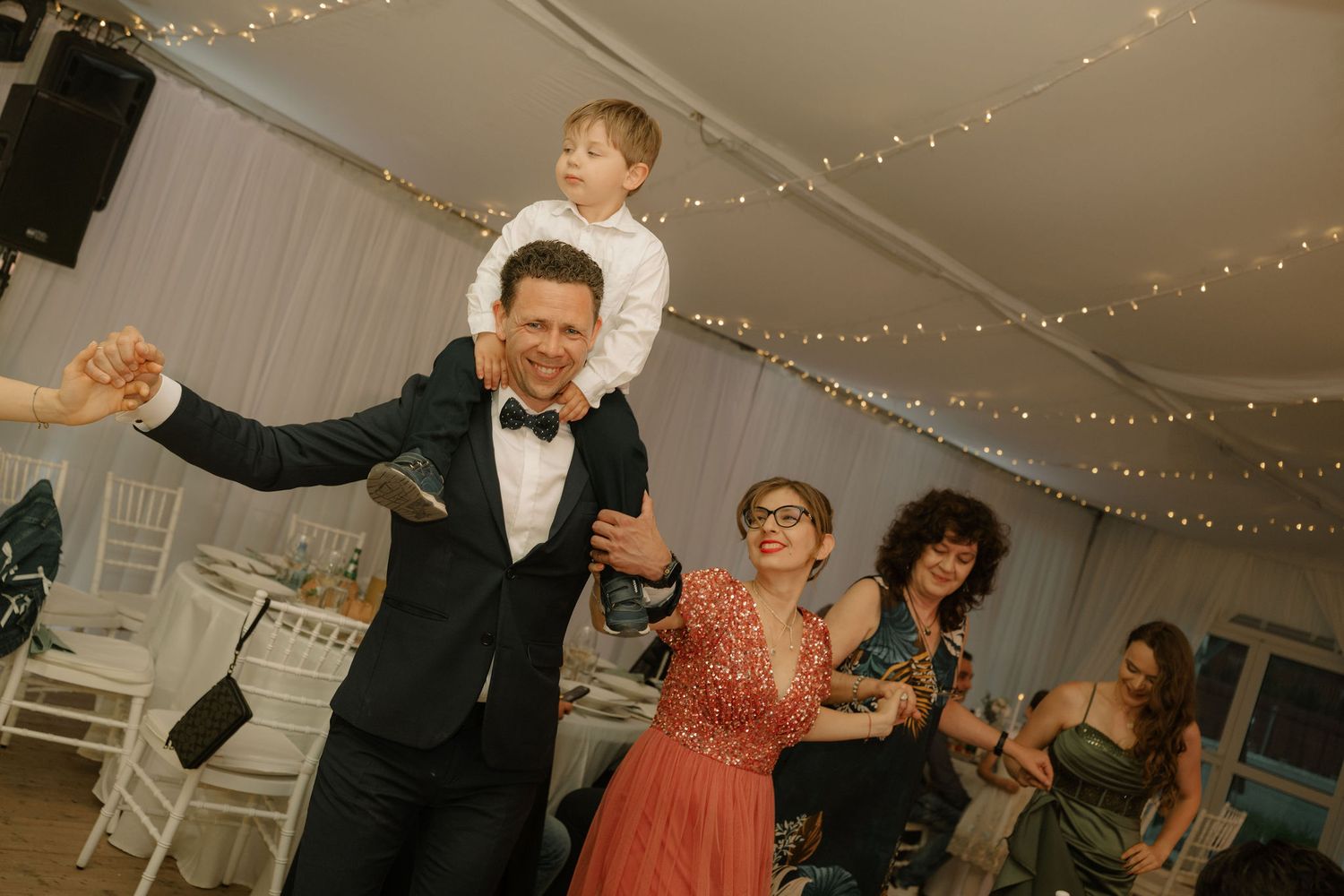 Man in tuxedo holds child on shoulders while dancing at elegant wedding reception with fairy lights.