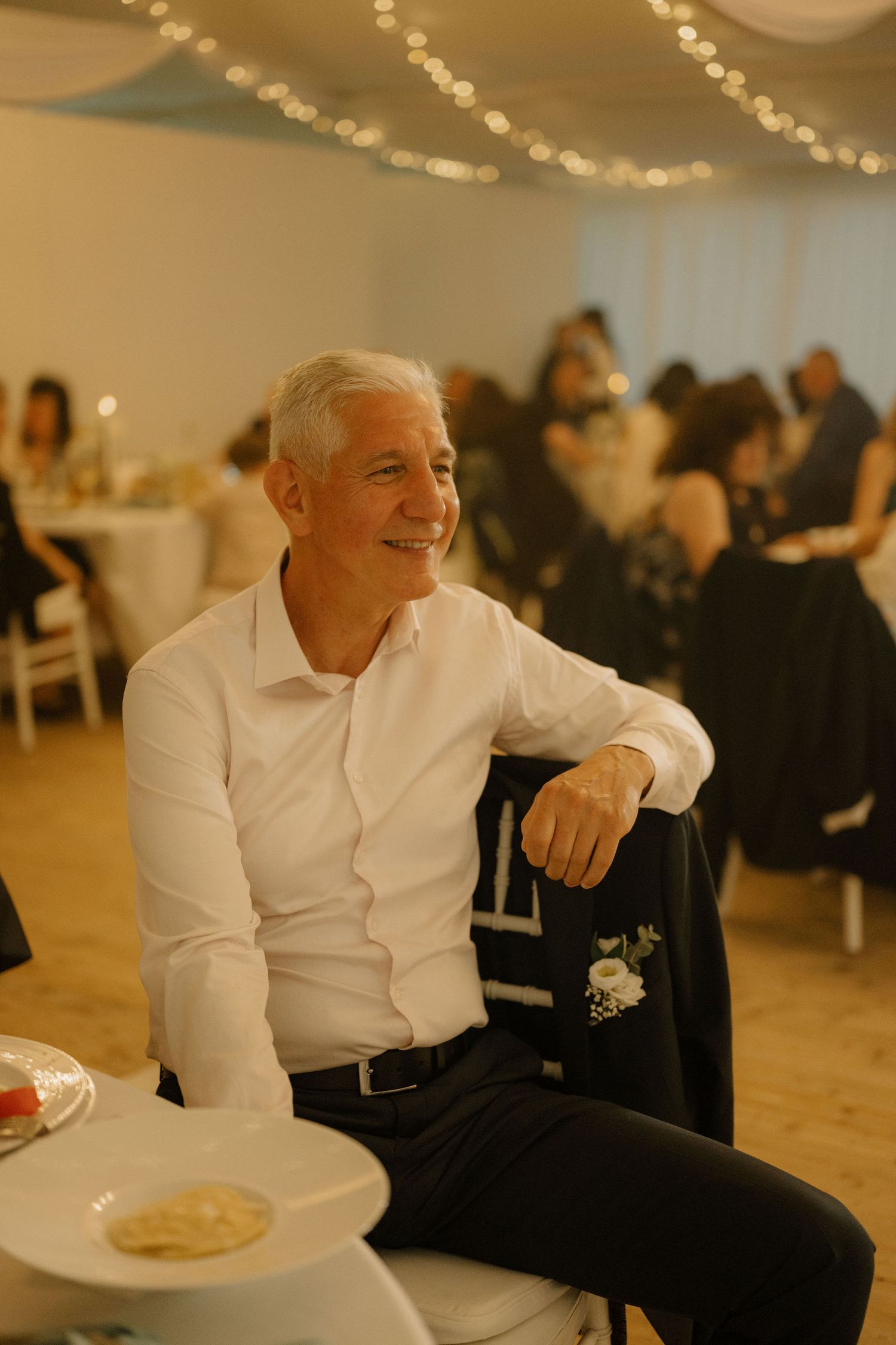 Smiling older man with white hair sits relaxed at an elegant wedding reception, wearing a white shirt and dark trousers.
