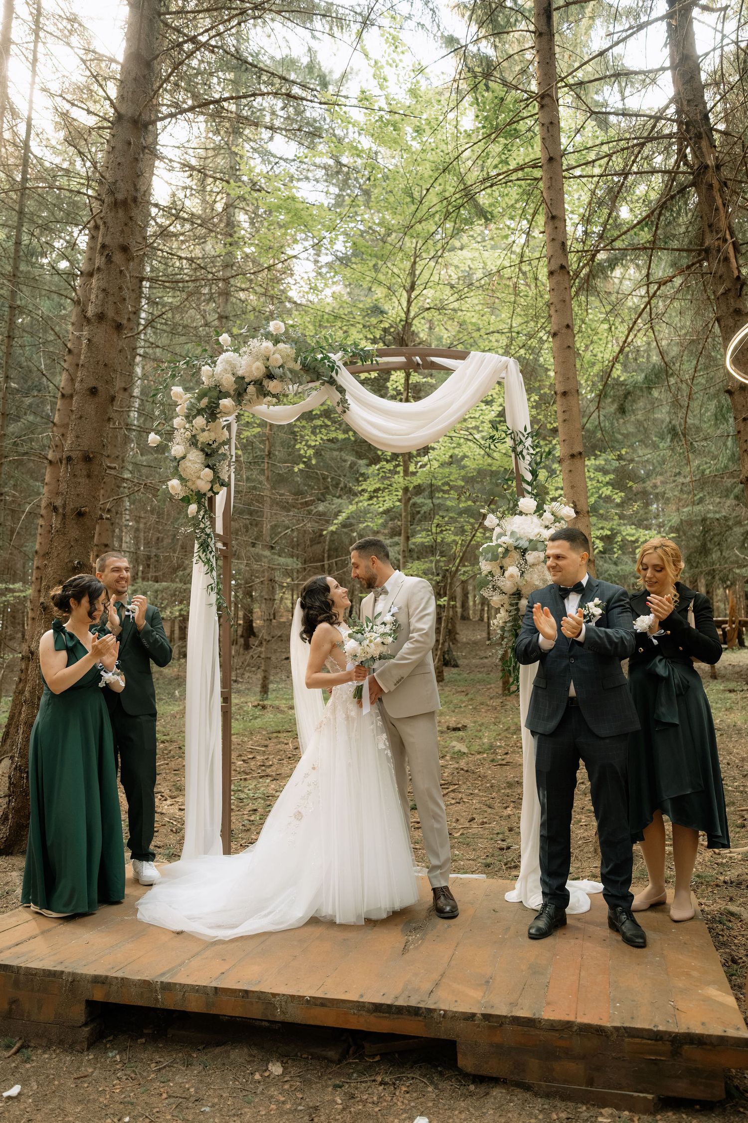 Bride and groom kissing under floral arch in forest wedding ceremony with bridesmaids in green dresses and guests applauding.