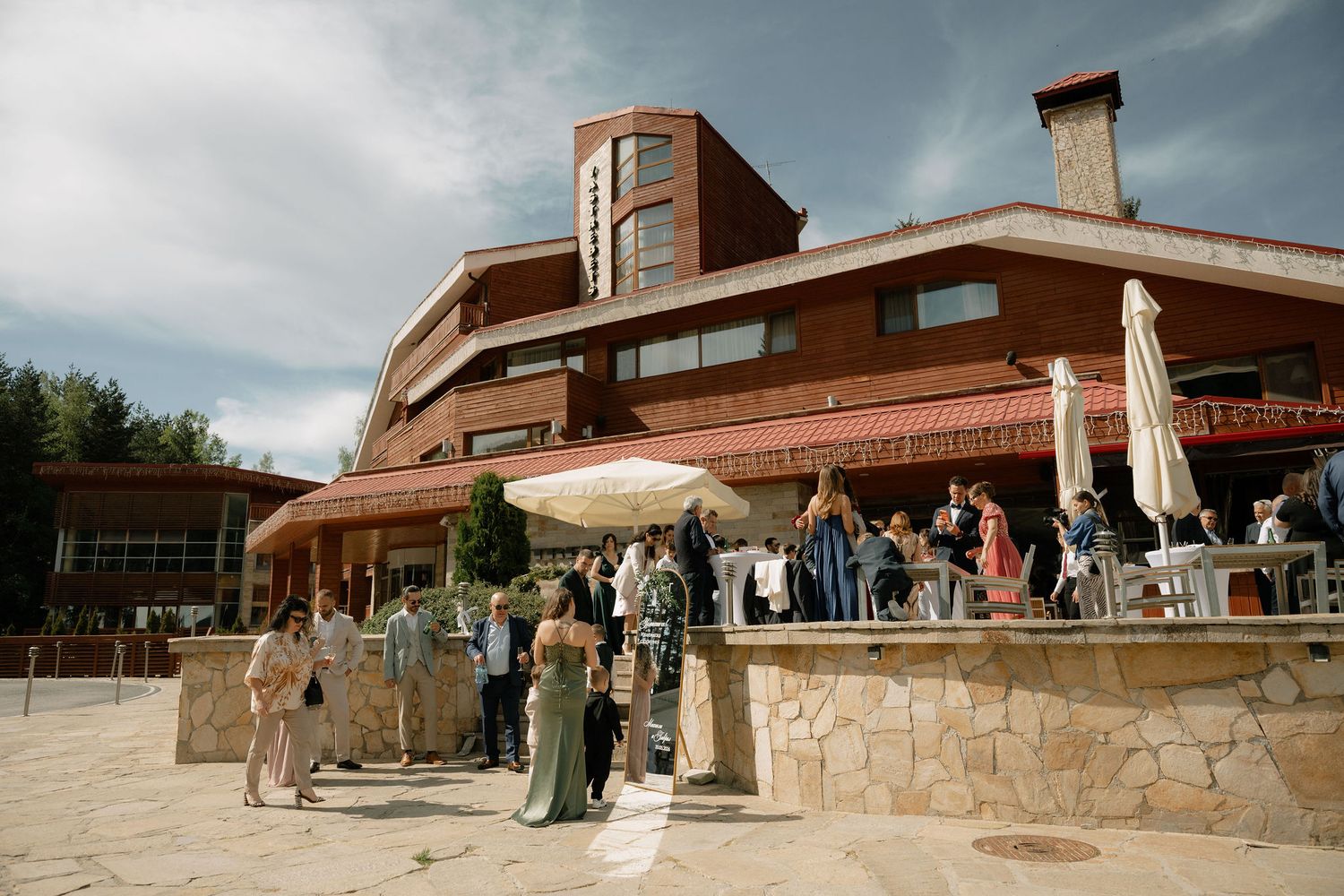 Guests gather outside a rustic brick venue with umbrellas and a stone terrace under a cloudy sky.