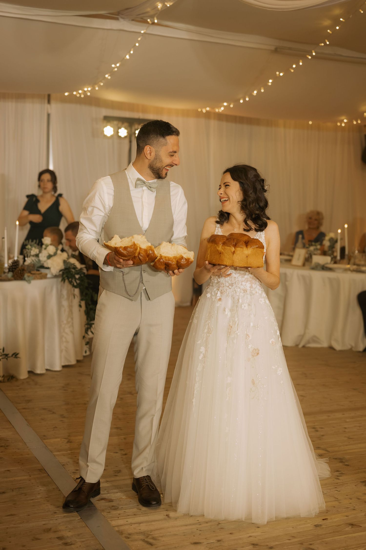 Smiling bride and groom holding decorative bread loaves at their elegant wedding reception with fairy lights.