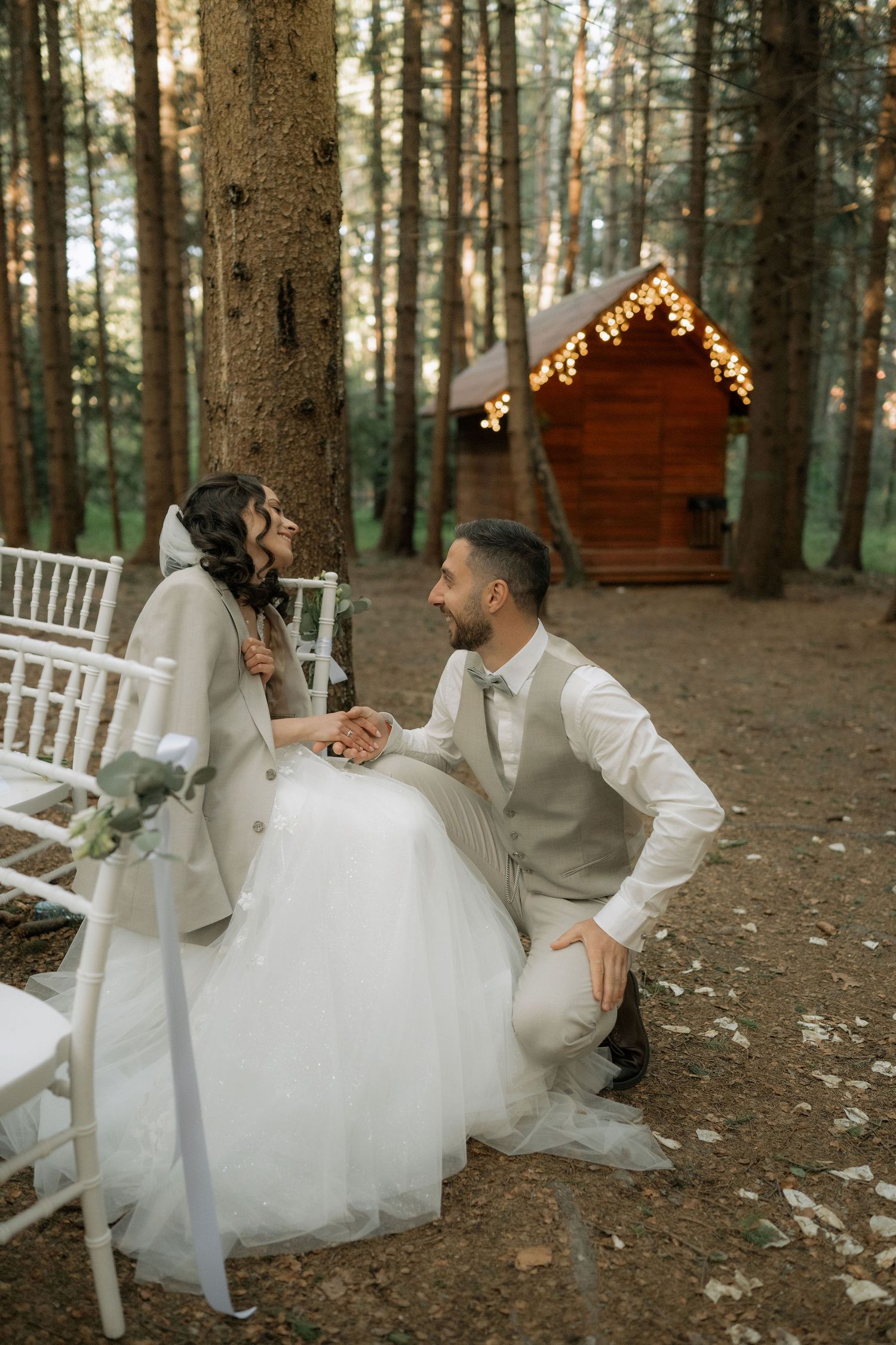 Bride and groom share a romantic moment in a forest wedding setting with fairy lights and wooden chapel backdrop.