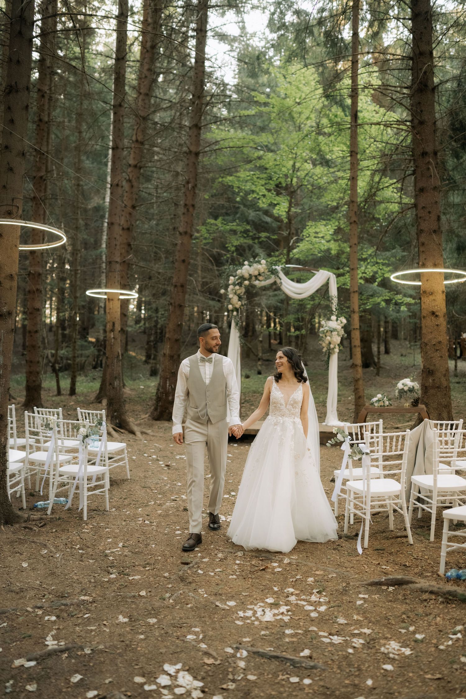 Bride and groom walk hand-in-hand down forest aisle with white chairs and floral arch backdrop.
