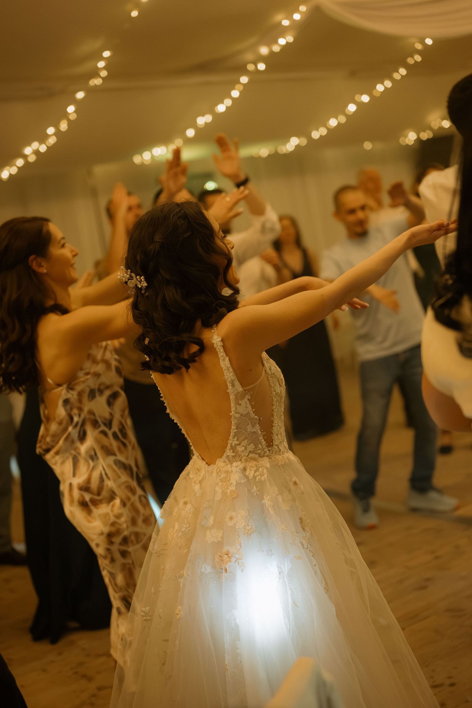 Bride in backless lace ball gown dancing with guests at elegant wedding reception with fairy lights.