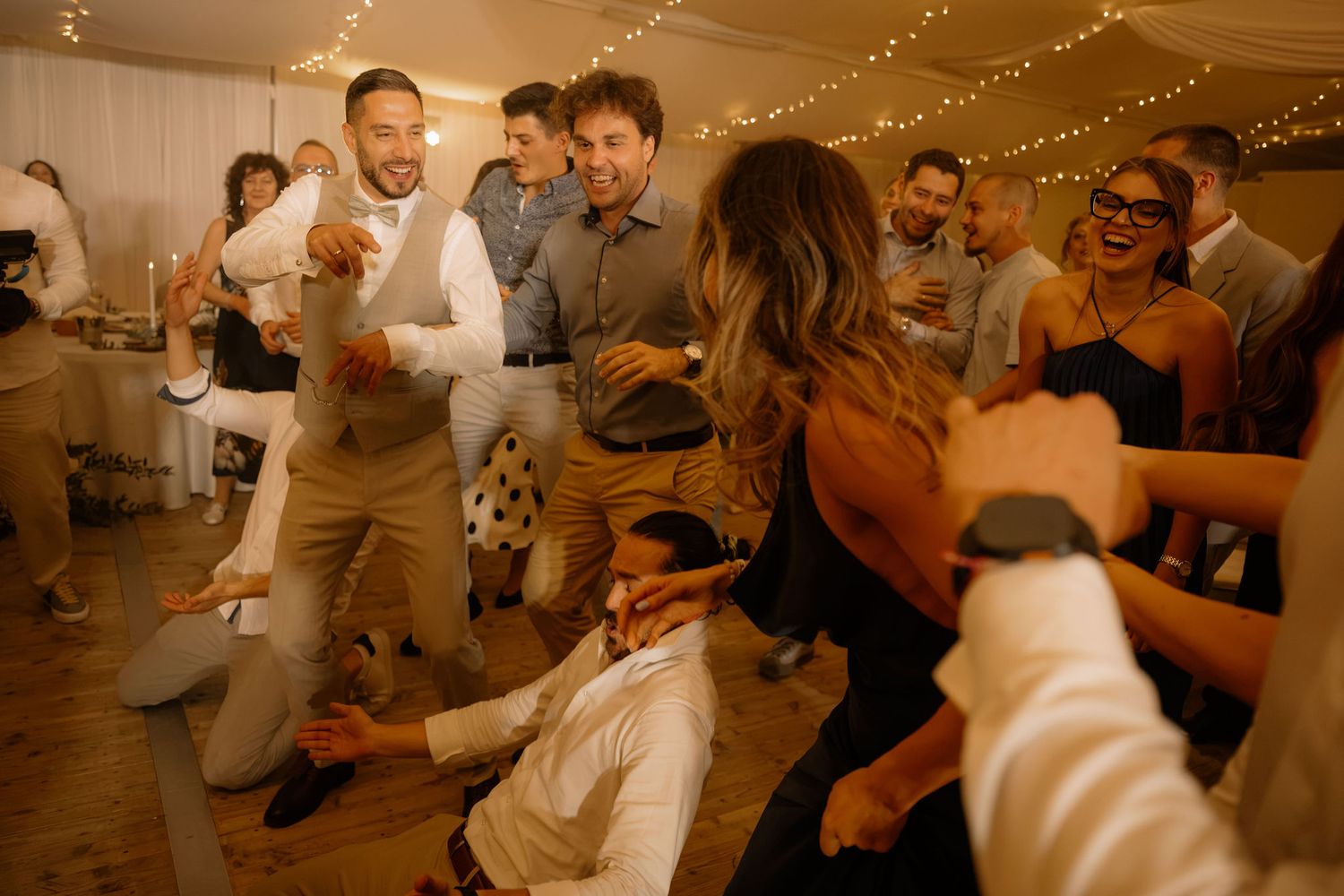 Guests dancing and celebrating at a lively wedding reception party under warm string lights.