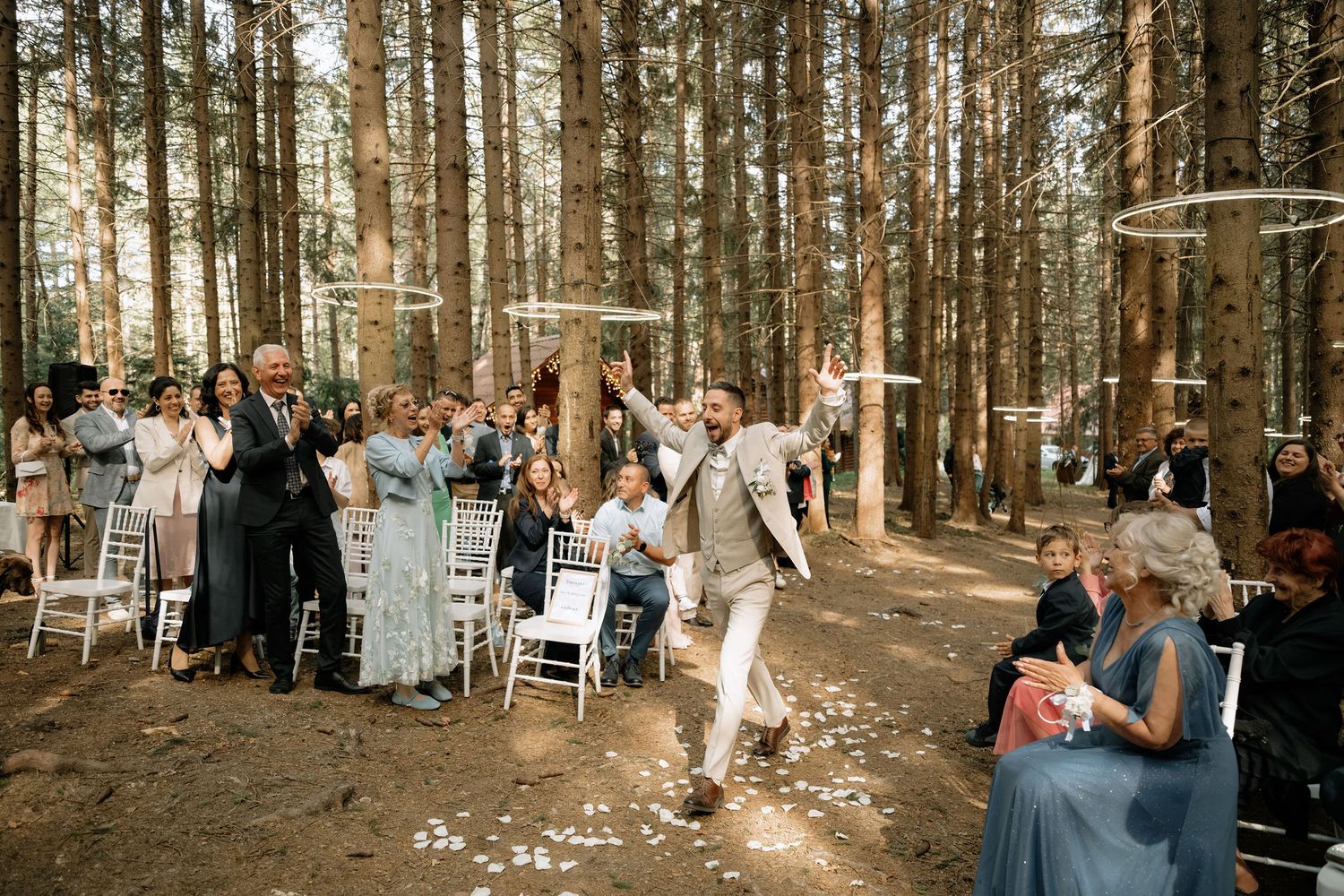 Groom celebrating joyfully at an outdoor forest wedding ceremony with guests seated on white chairs among tall pine trees.