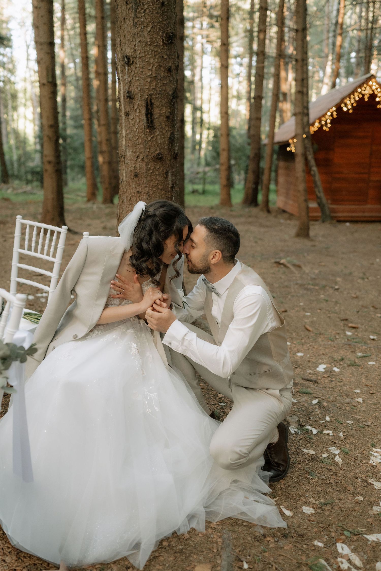 Bride and groom share a romantic kiss in a pine forest, with the groom kneeling beside his bride in a white gown.