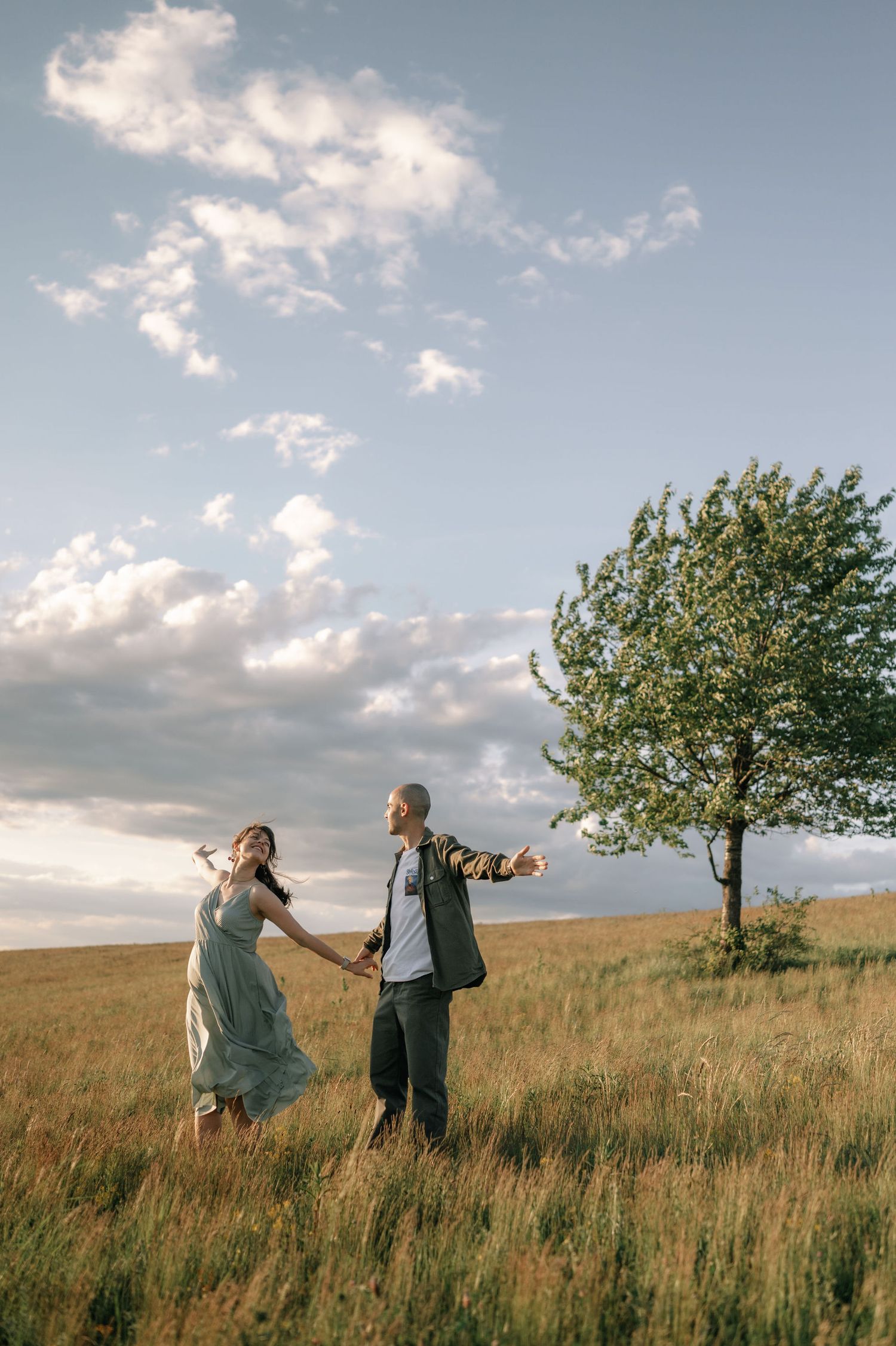 Couple holding hands gazing upward in a sunlit grassy field with a solitary tree under a cloudy sky.