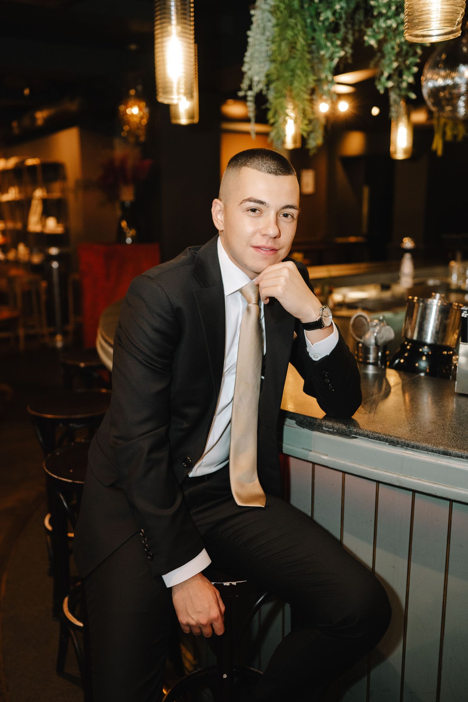 A person in a black suit sits at a modern bar counter with pendant lights and greenery hanging from above.
