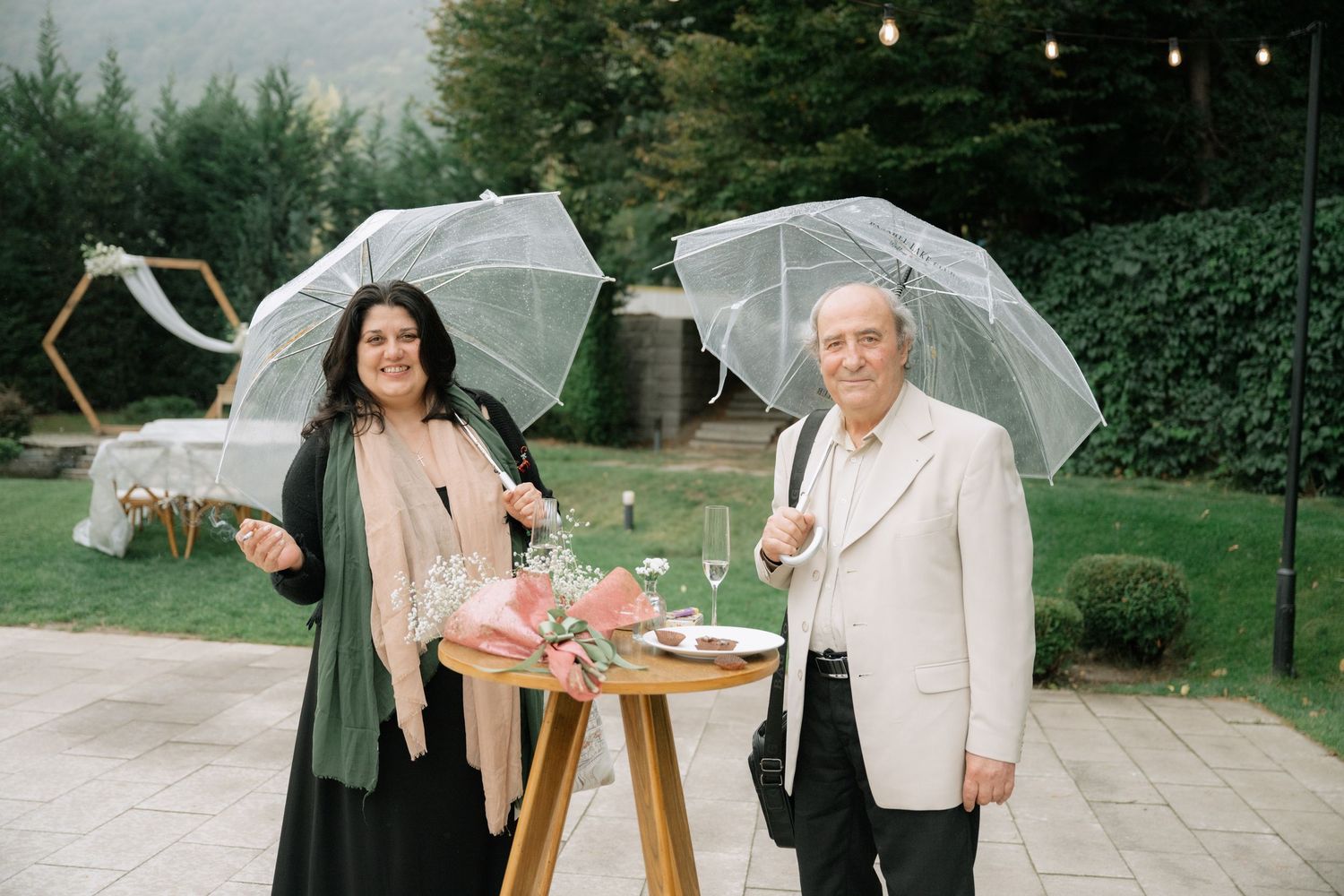 Two people stand with clear umbrellas next to a wooden table with flowers at an outdoor garden event.