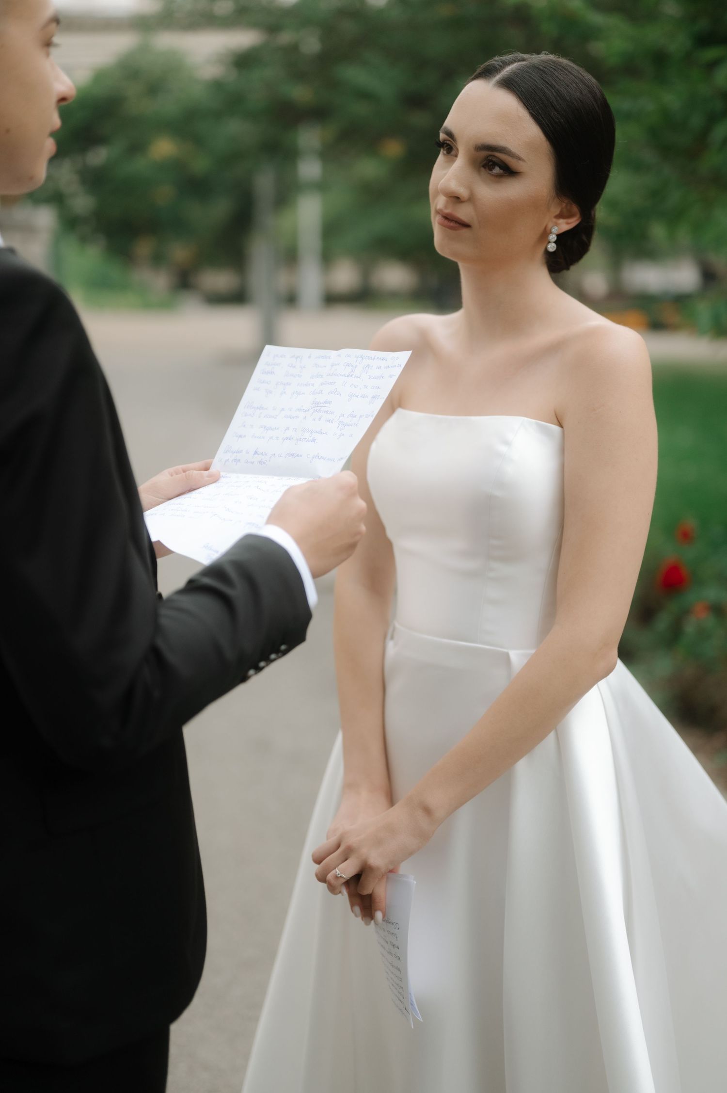 A couple exchanging handwritten vows during an outdoor wedding ceremony with greenery in the background.
