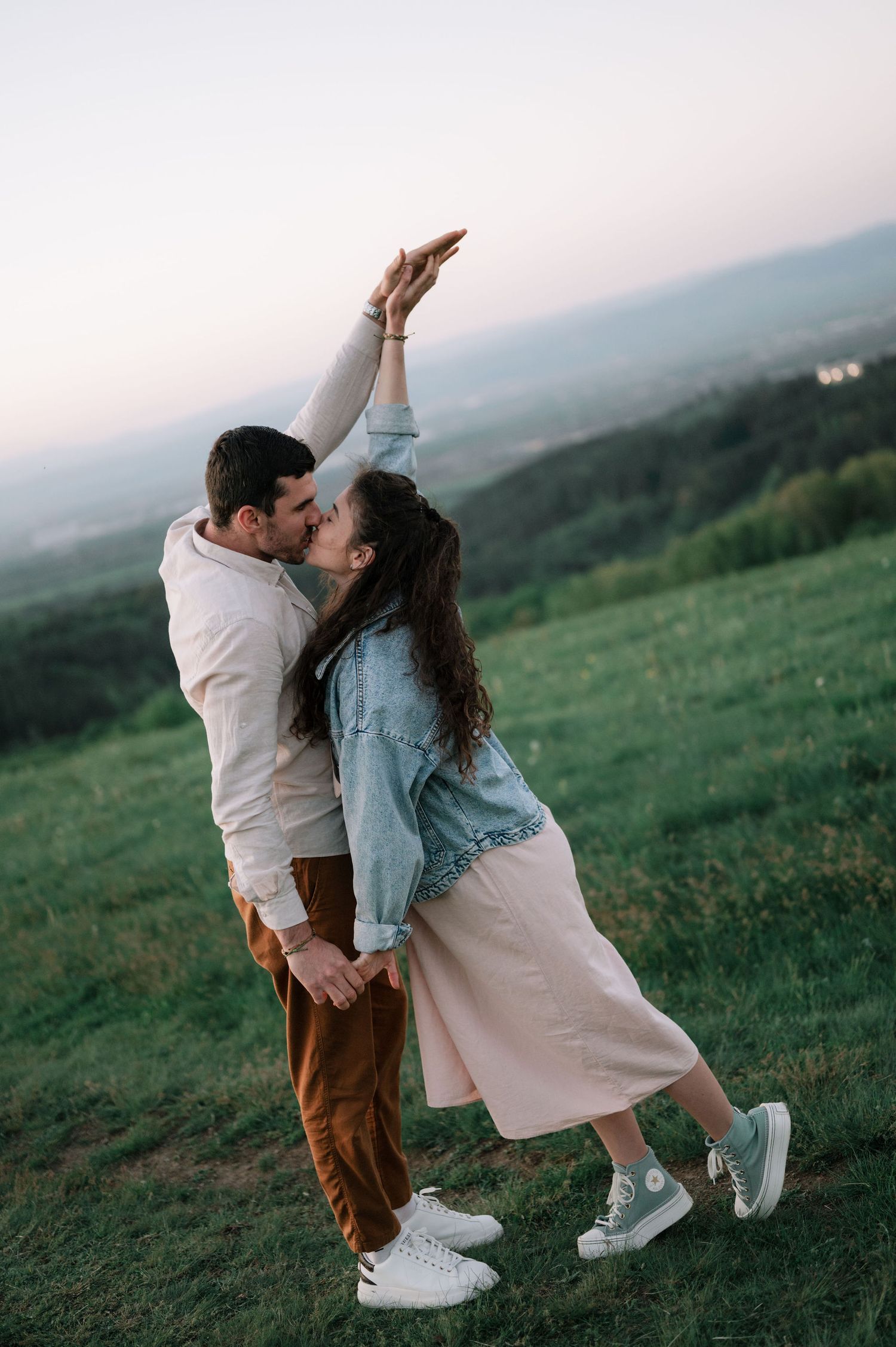 A couple shares a romantic kiss on a misty hillside while wearing casual white and brown outfits.
