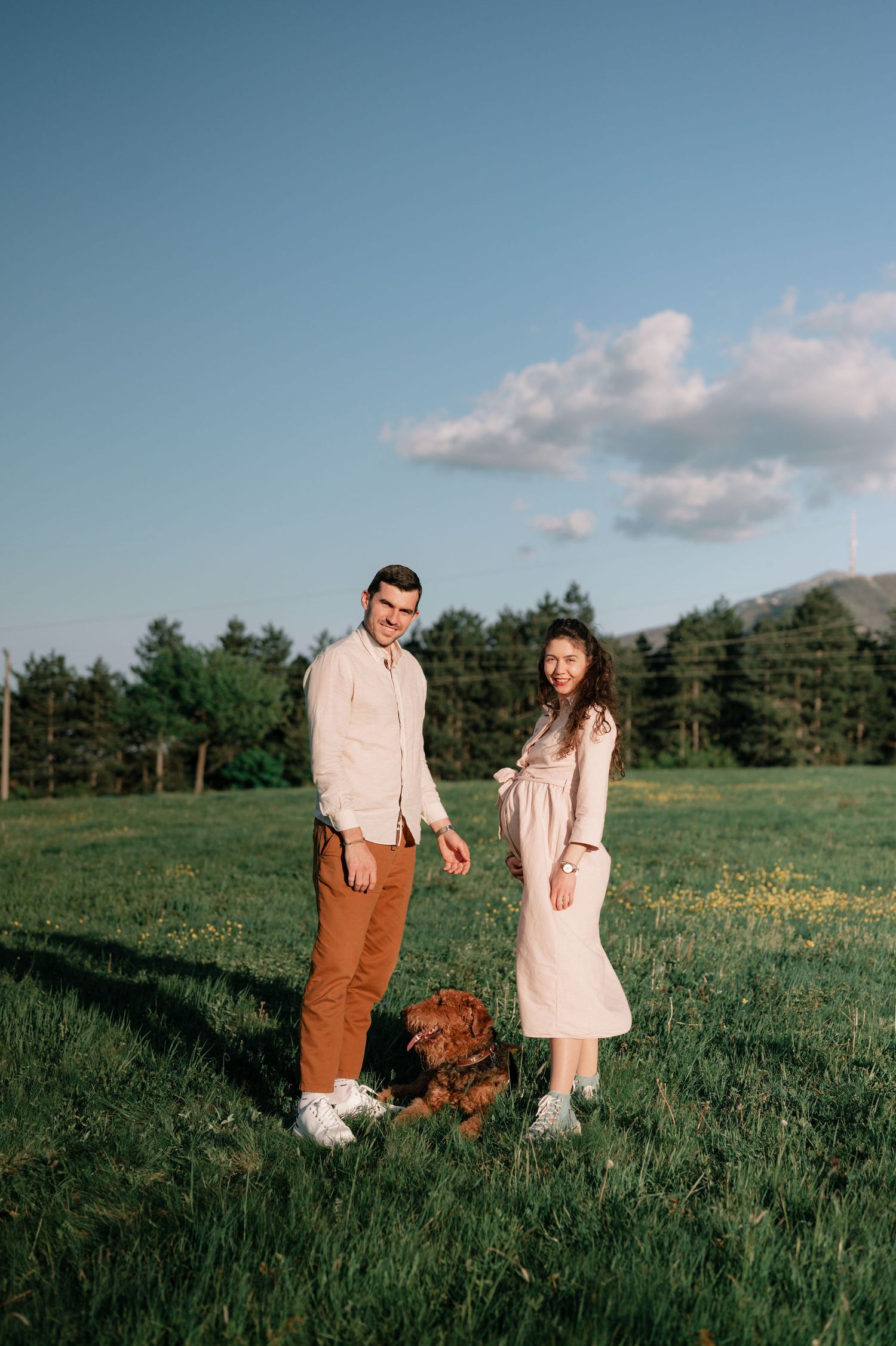 A couple stands in a green field with their dog on a sunny day with blue skies and white clouds in the background.