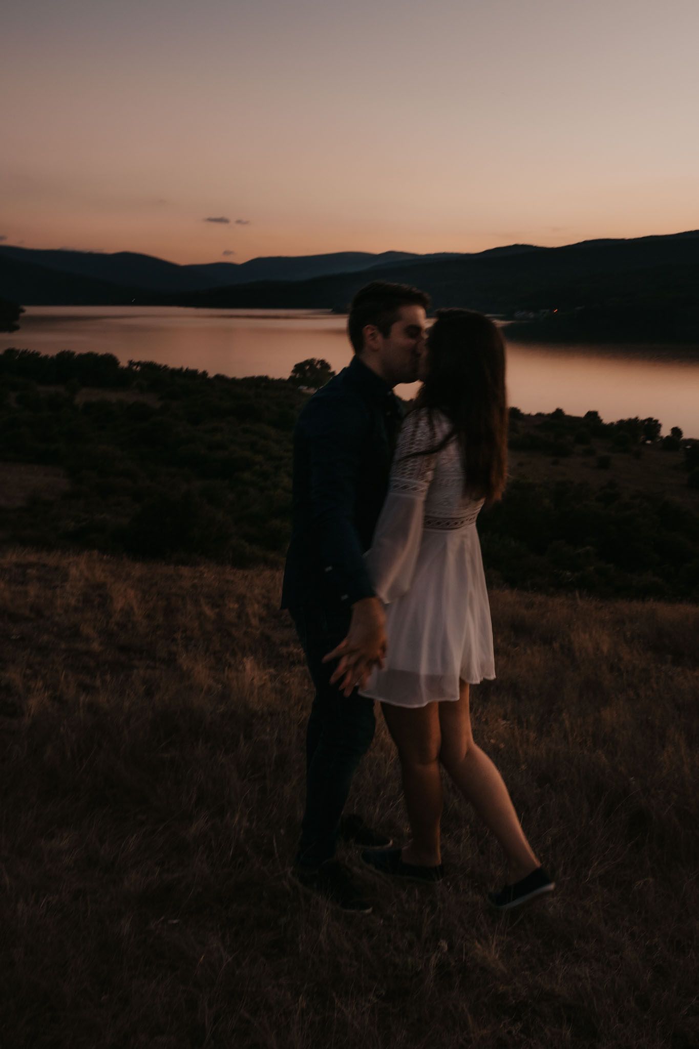 A romantic sunset silhouette of a couple embracing on a hilltop overlooking a serene lake.