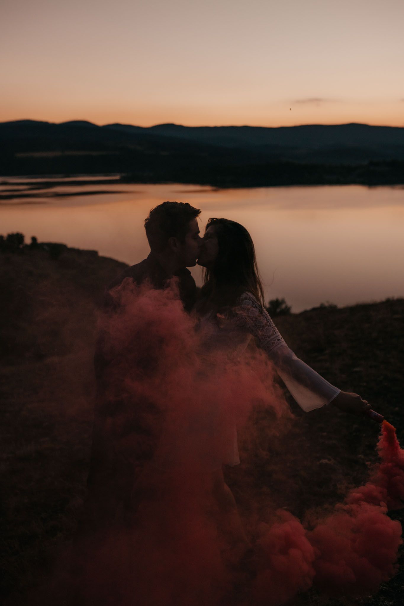 Silhouetted figures stand among red smoke clouds against a serene lakeside sunset backdrop.