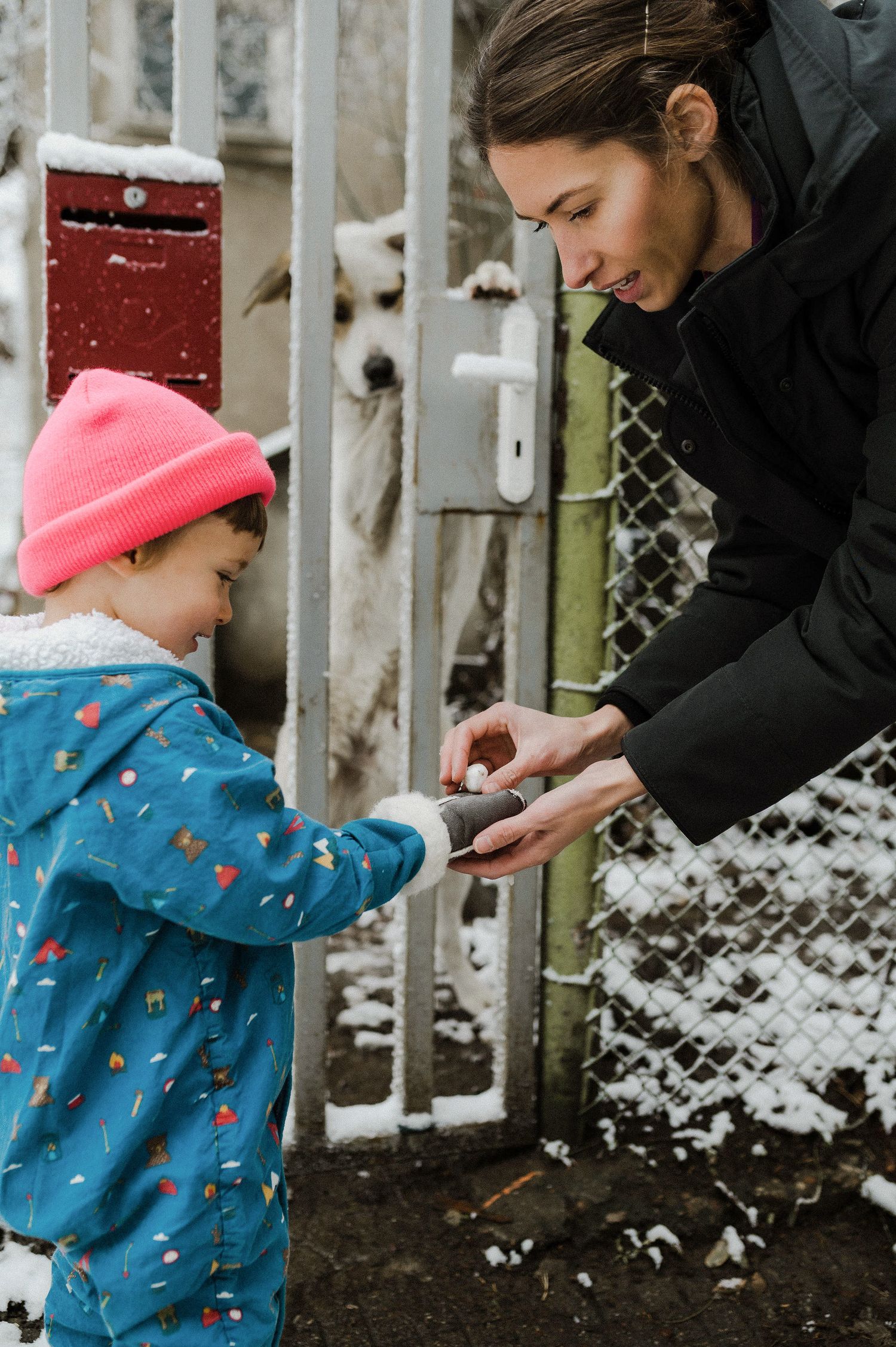 A child in a blue jacket and pink hat explores snow-covered ground while an adult assists in winter exploration.