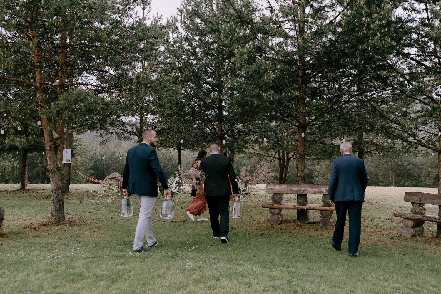 Small groups of guests walking across a grassy outdoor venue with pine trees.
