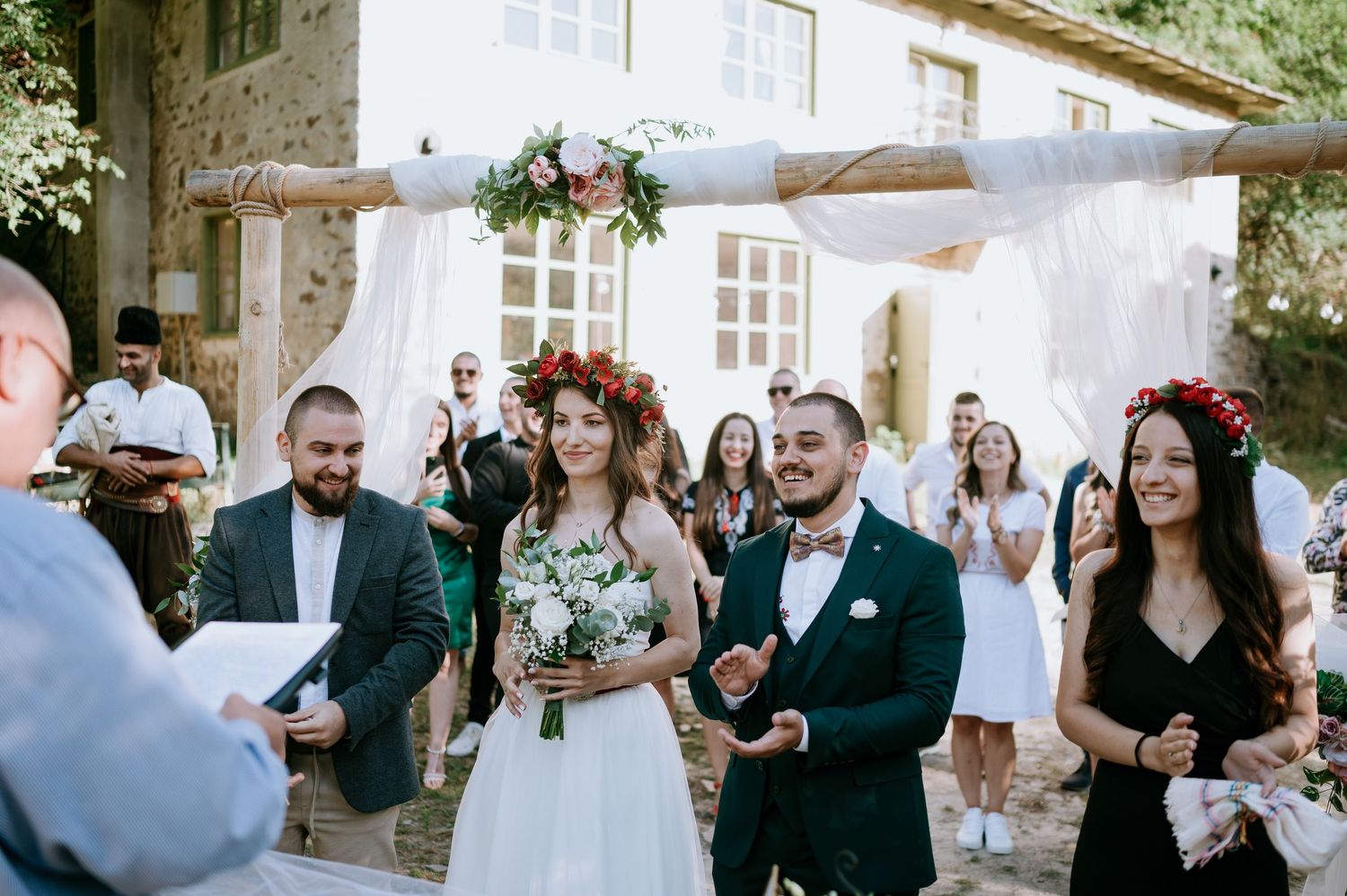 Wedding guests gather outdoors at a rustic venue with white curtains and floral decorations.
