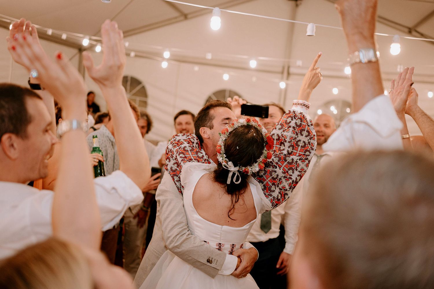 Wedding guests dance and celebrate under string lights in a white tent reception venue.