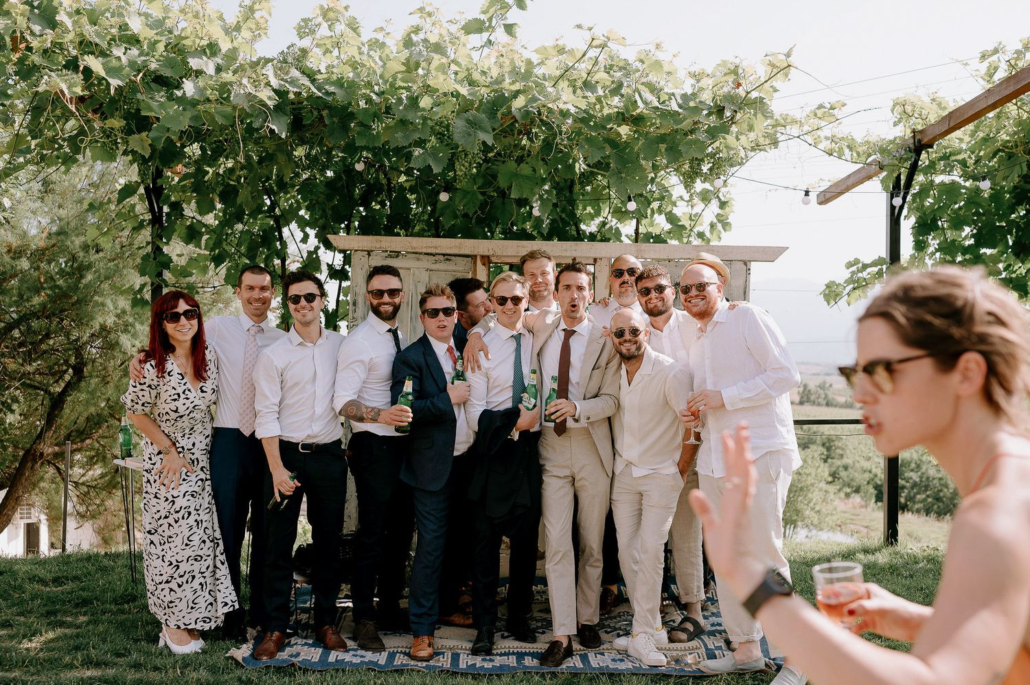 Wedding guests gather for a group photo under a leafy green arbor.