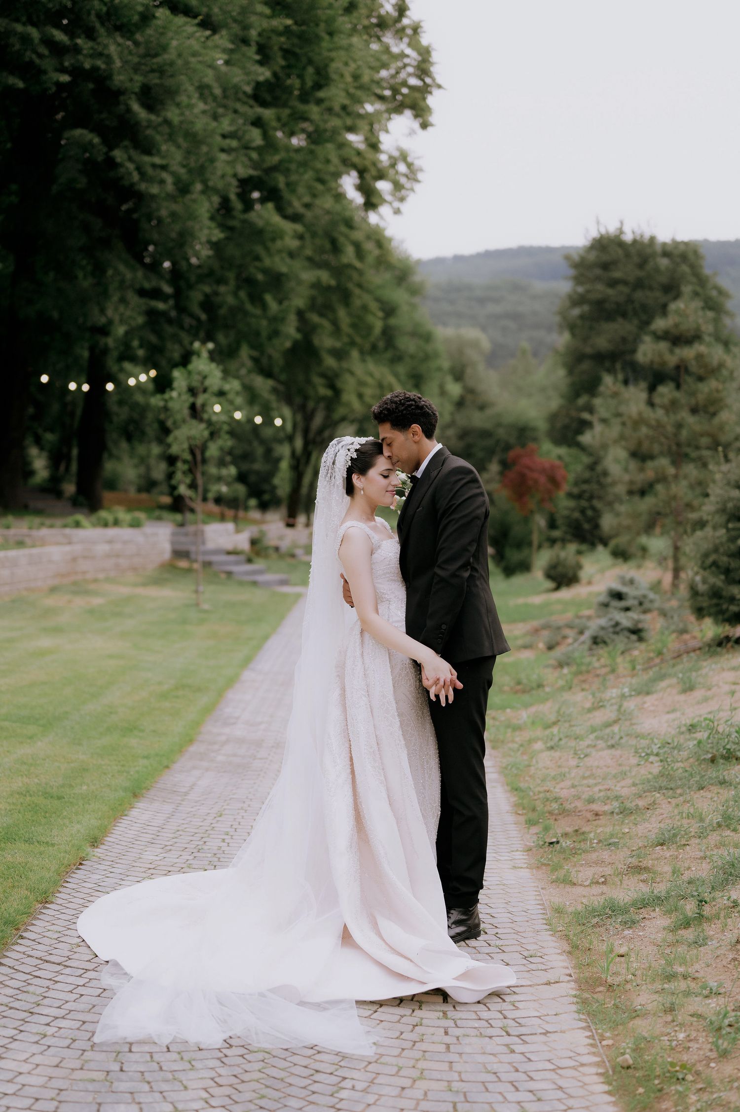 A romantic wedding couple shares intimate moments on a stone pathway surrounded by lush greenery.