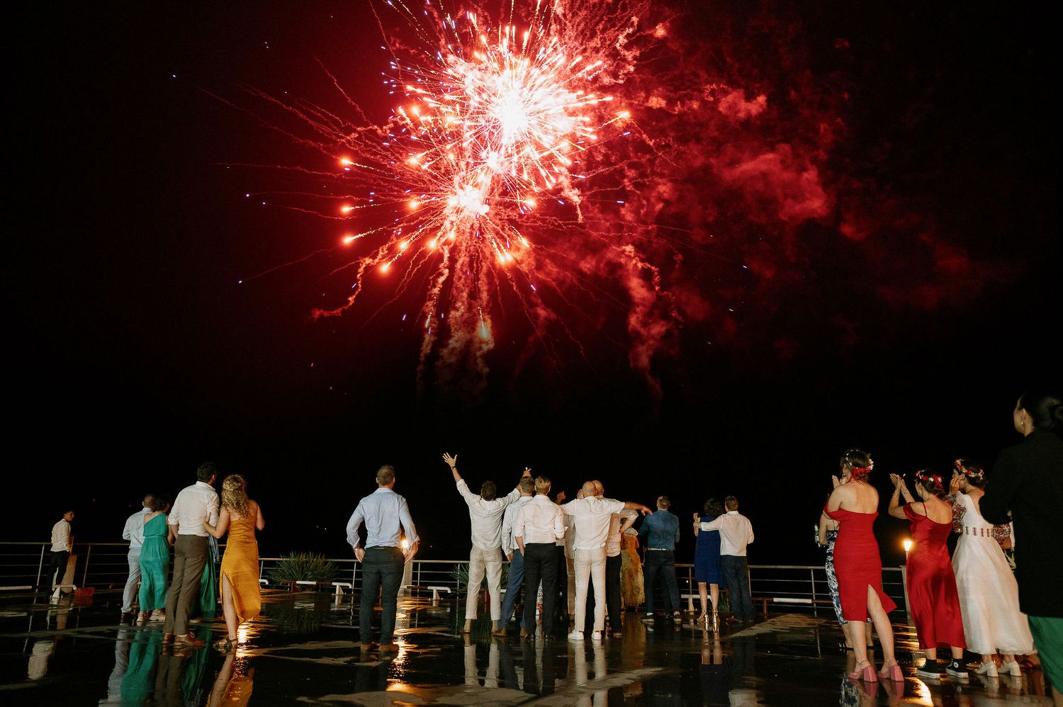 Red fireworks illuminate the night sky as wedding guests watch from below.