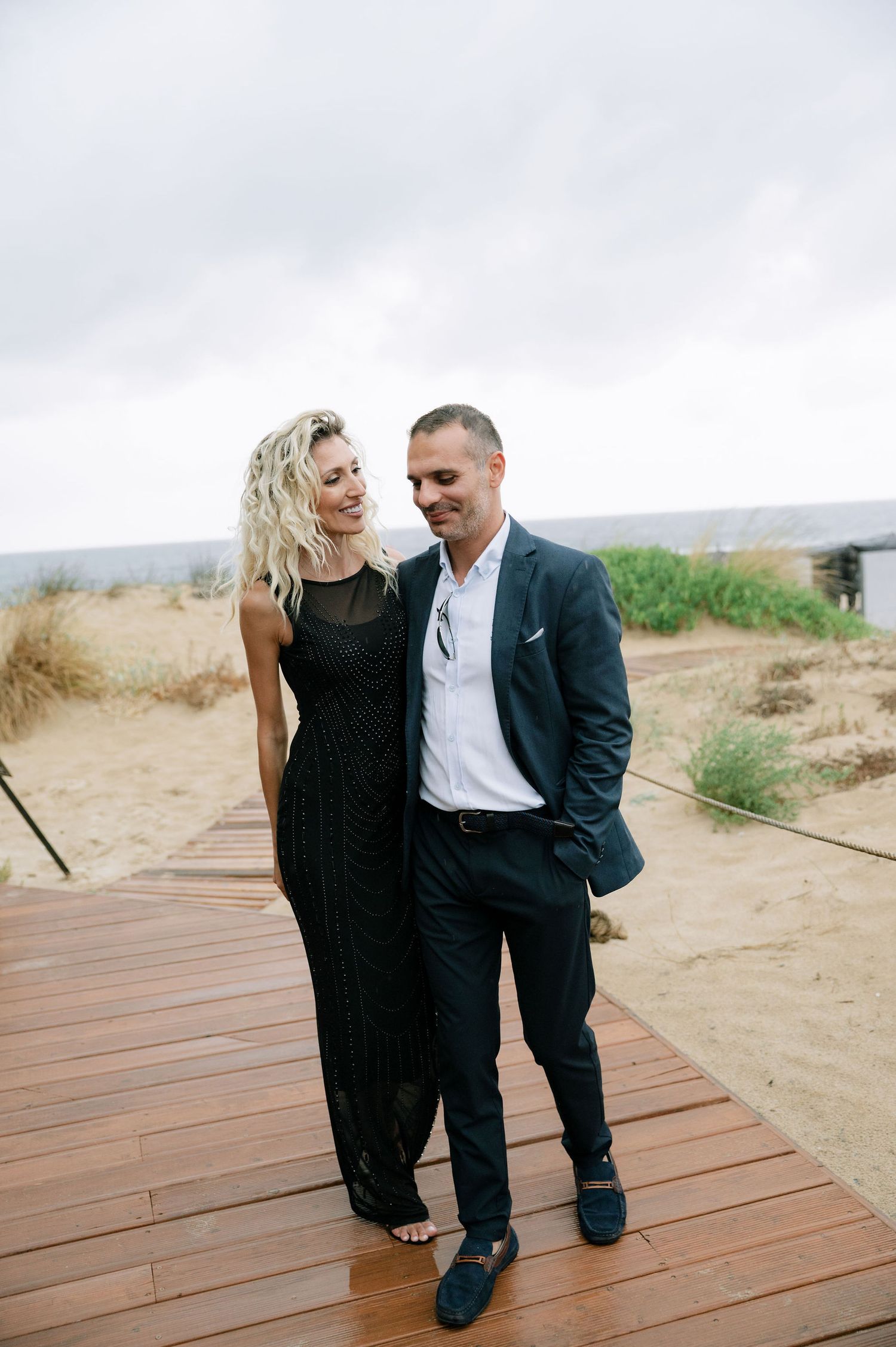 Couple in formal attire walking along wooden boardwalk on beach.