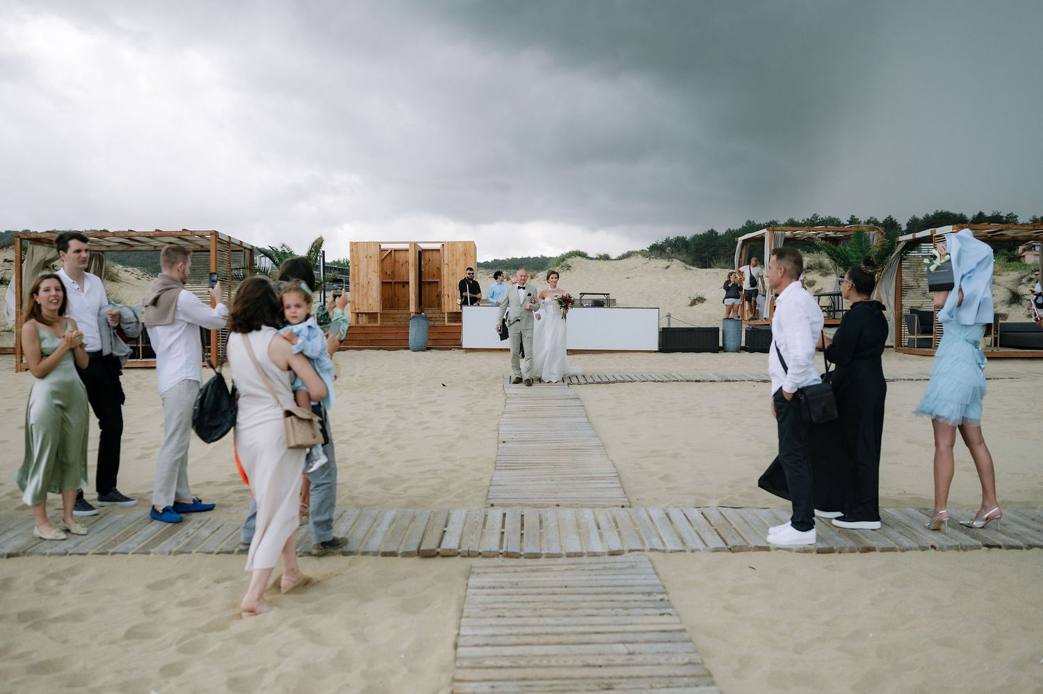 Wooden boardwalk stretches across beach sand with wedding guests gathered for celebration.