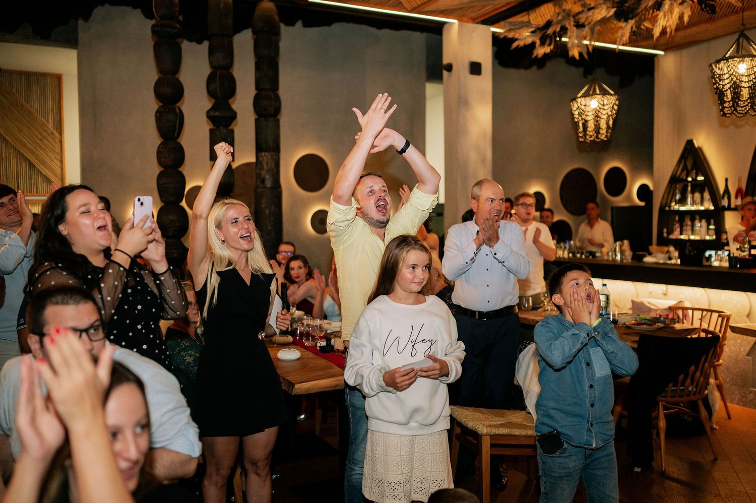 Wedding guests cheer and celebrate during a reception in a rustic venue with hanging lanterns.