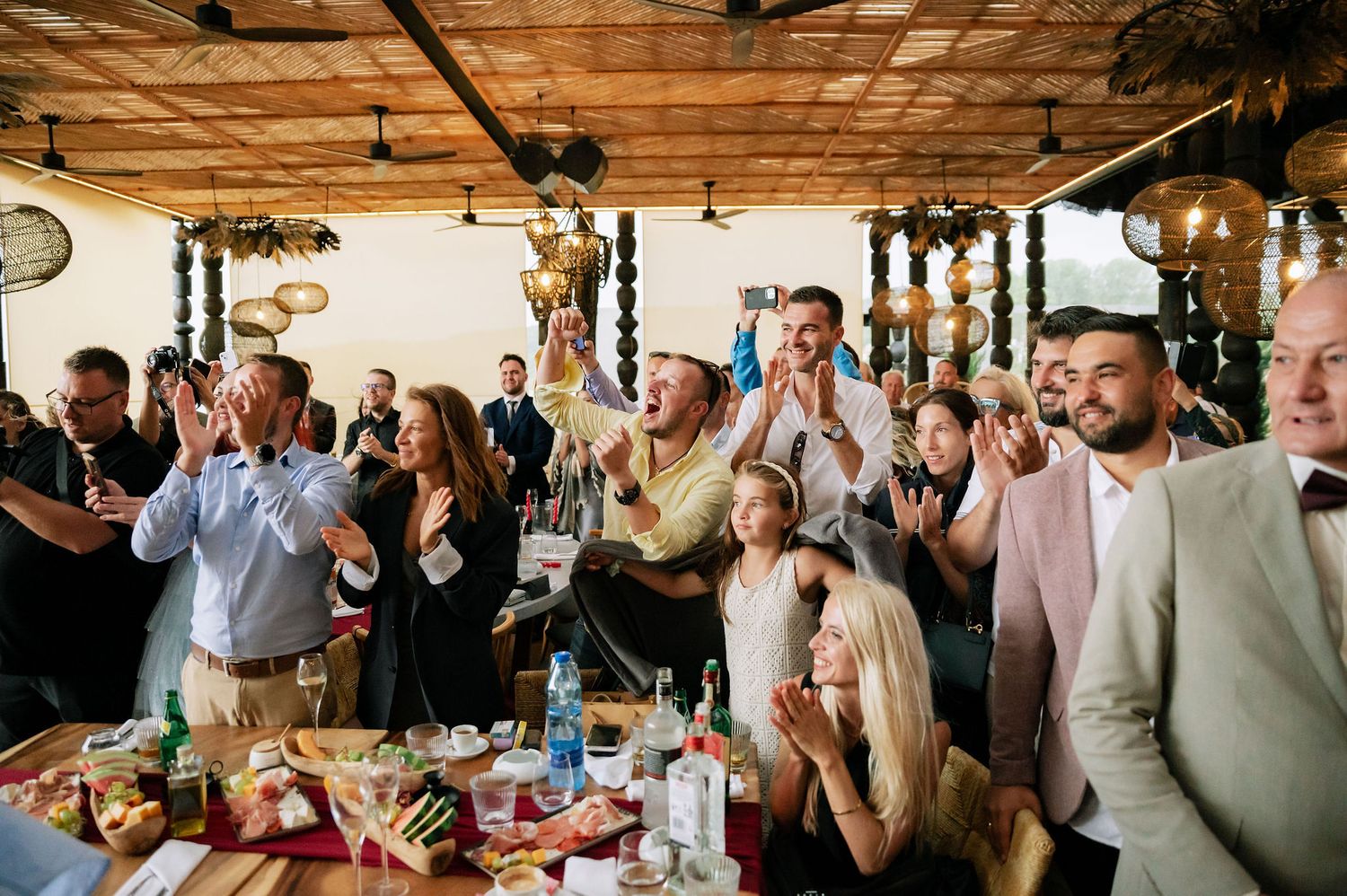 Wedding celebration with guests clapping and cheering in a rustic indoor venue with wooden beams.