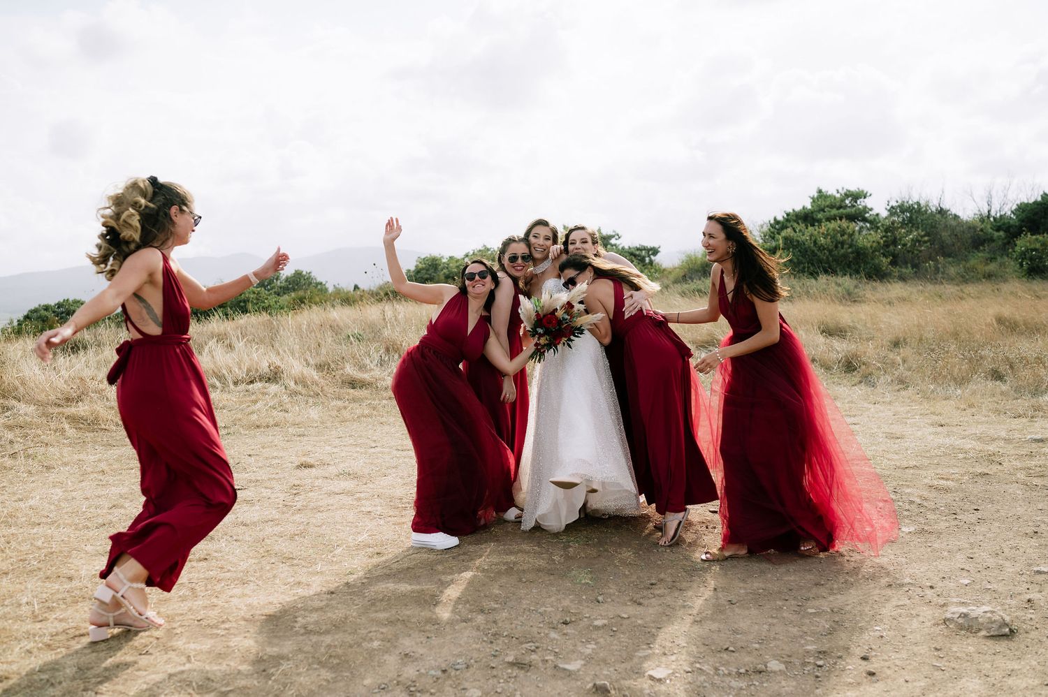 Bridal party in red dresses sits together on grass for casual photo.
