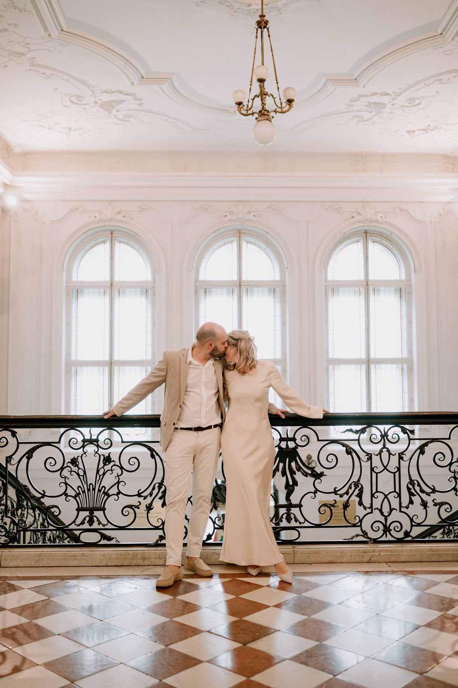 A couple poses together in a grand ballroom with arched windows and ornate ceiling details.