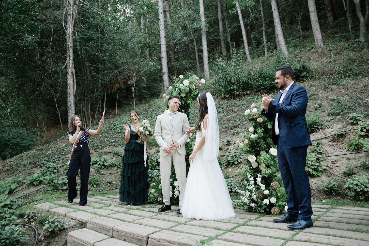 Outdoor woodland wedding ceremony takes place on a wooden deck surrounded by trees.