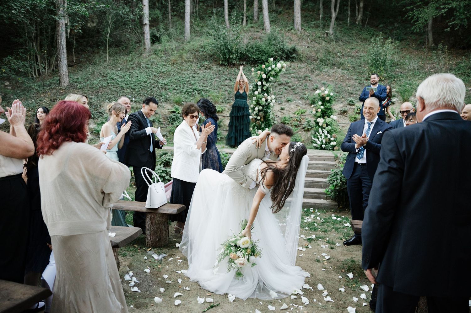 Wedding ceremony guests gather in a wooded outdoor setting with scattered white flower petals.