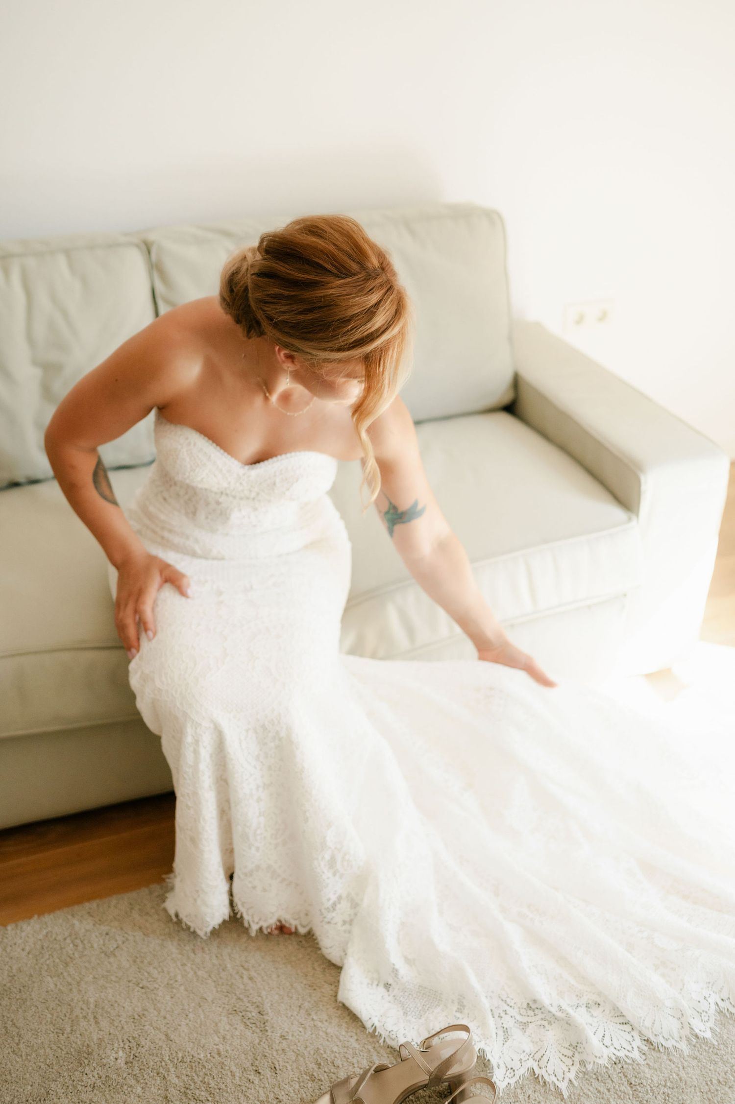 A bride in a lace wedding gown adjusts her dress while sitting on a white couch.