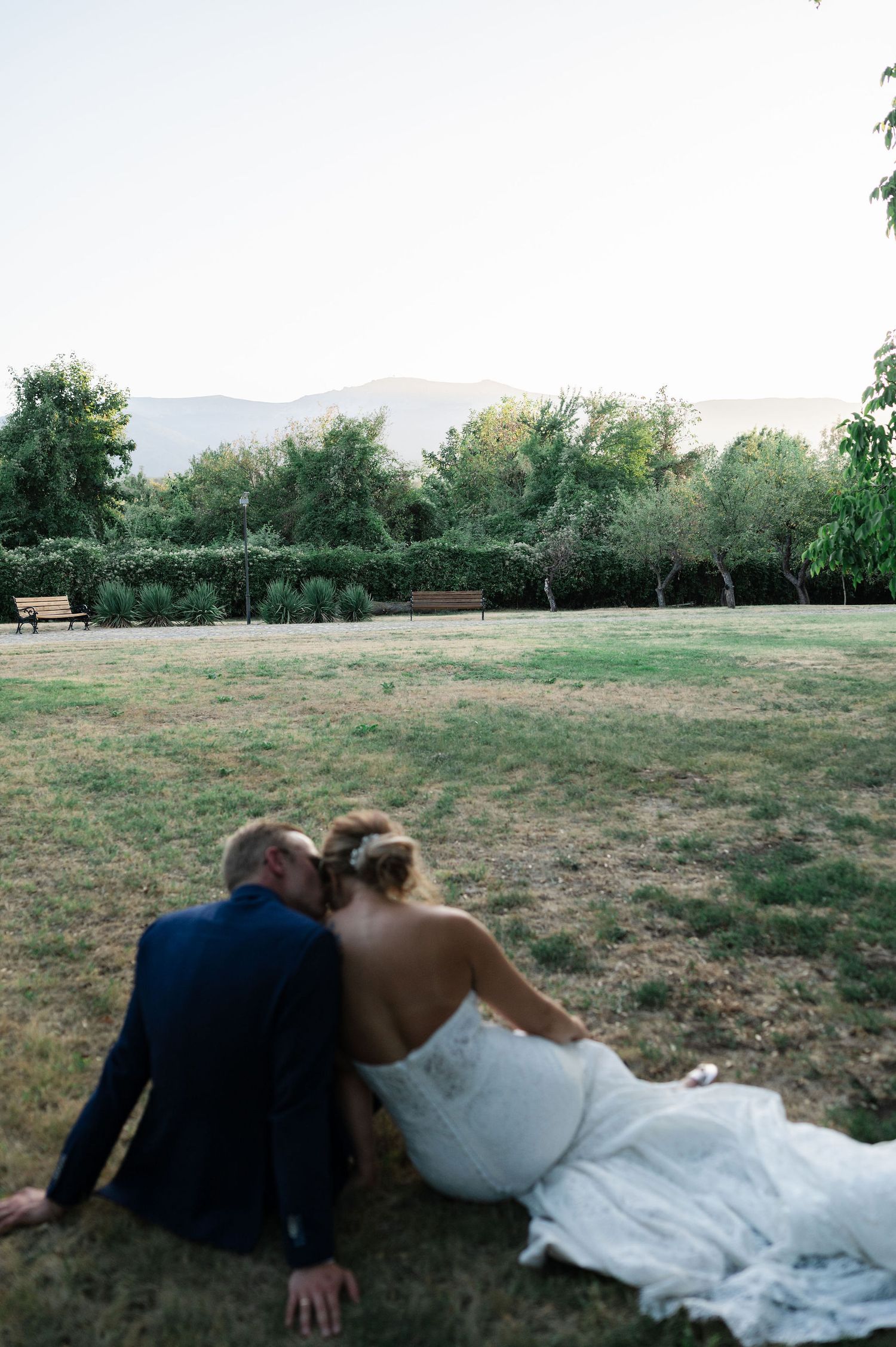 Couple in wedding attire lying together on grassy field with mountains in background.