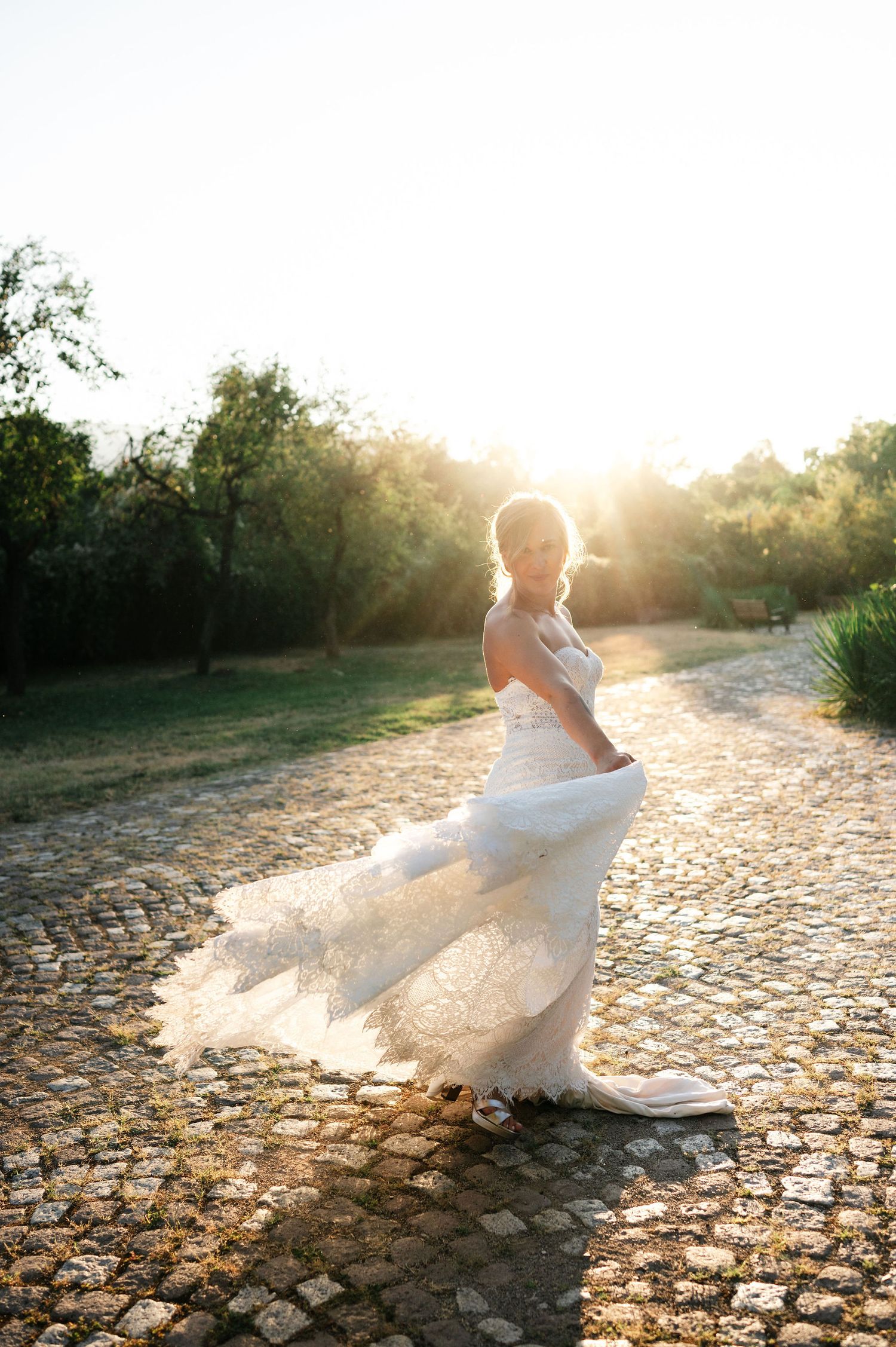 Bride spins in white lace gown on cobblestone path at sunset.