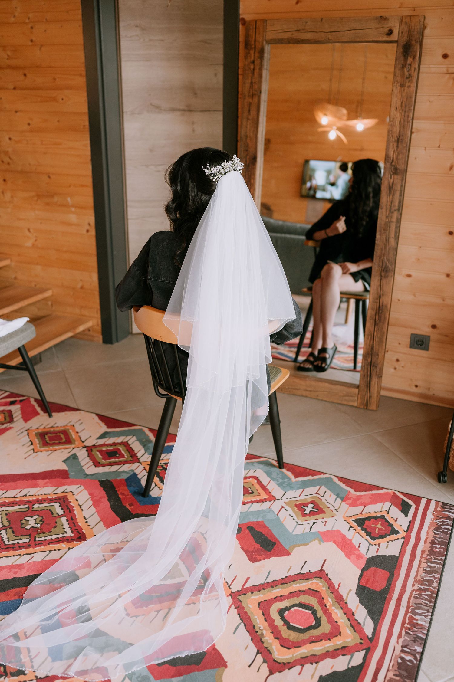 Wedding dress with flowing veil draped across a colorful geometric patterned rug in a wooden cabin.