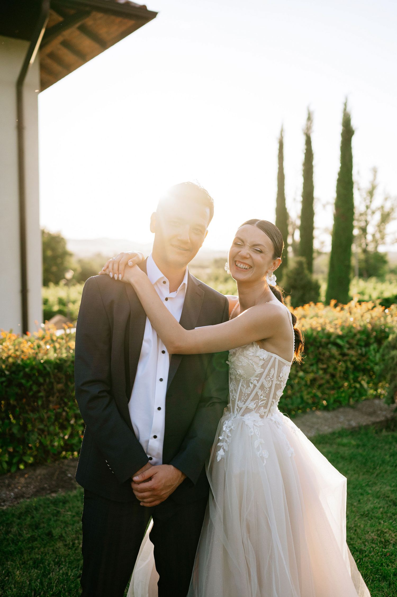 A romantic sunset wedding photo showing a couple dancing together in formal attire on a lawn with cypress trees.