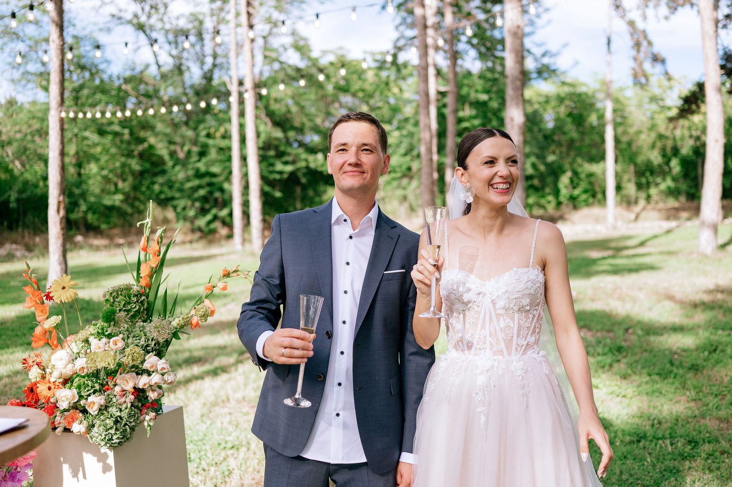 Wedding couple shares champagne in a sunlit forest setting with string lights and decorative flowers.